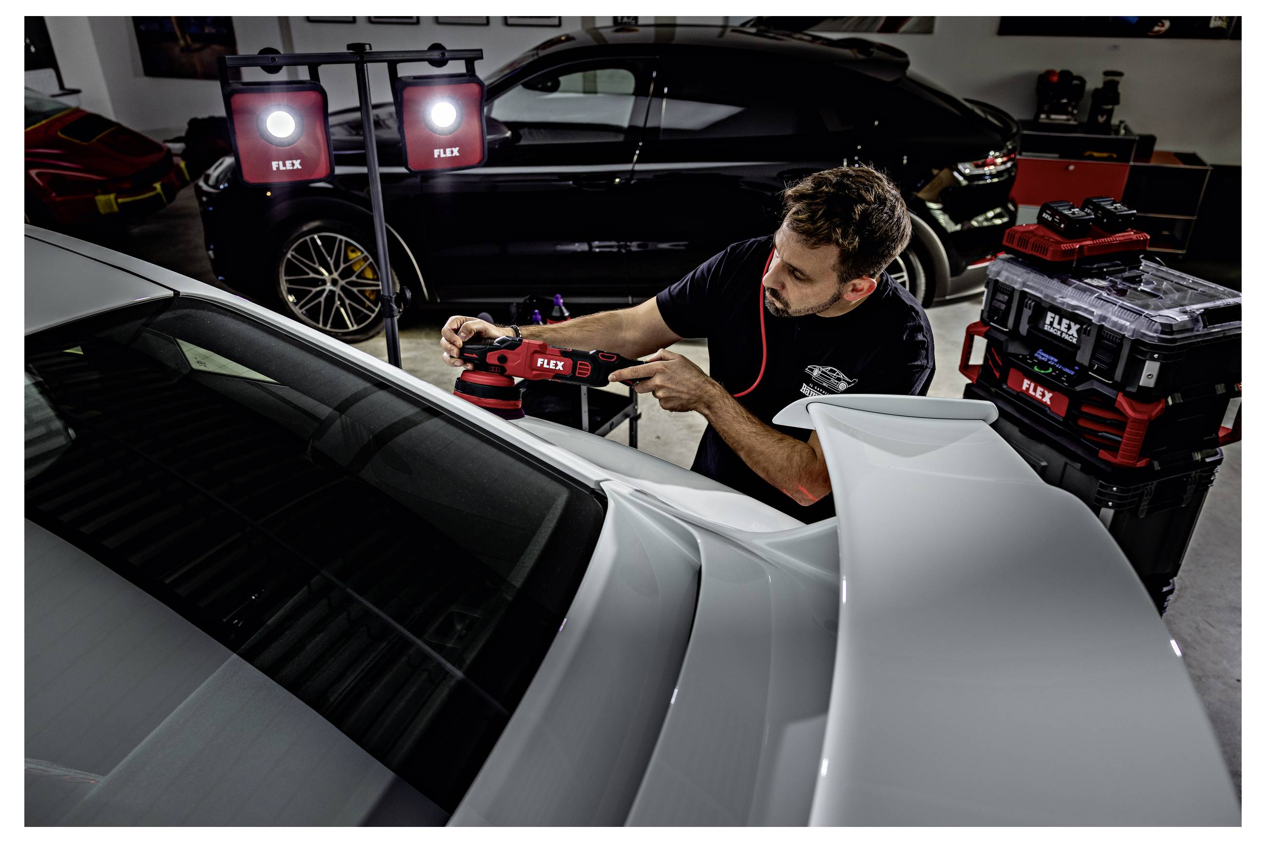 Mechanic leští bílý sportovní vůz s FLEX leštičkou v garáži, v pozadí je černý vůz a naskládané kufry s nářadím.