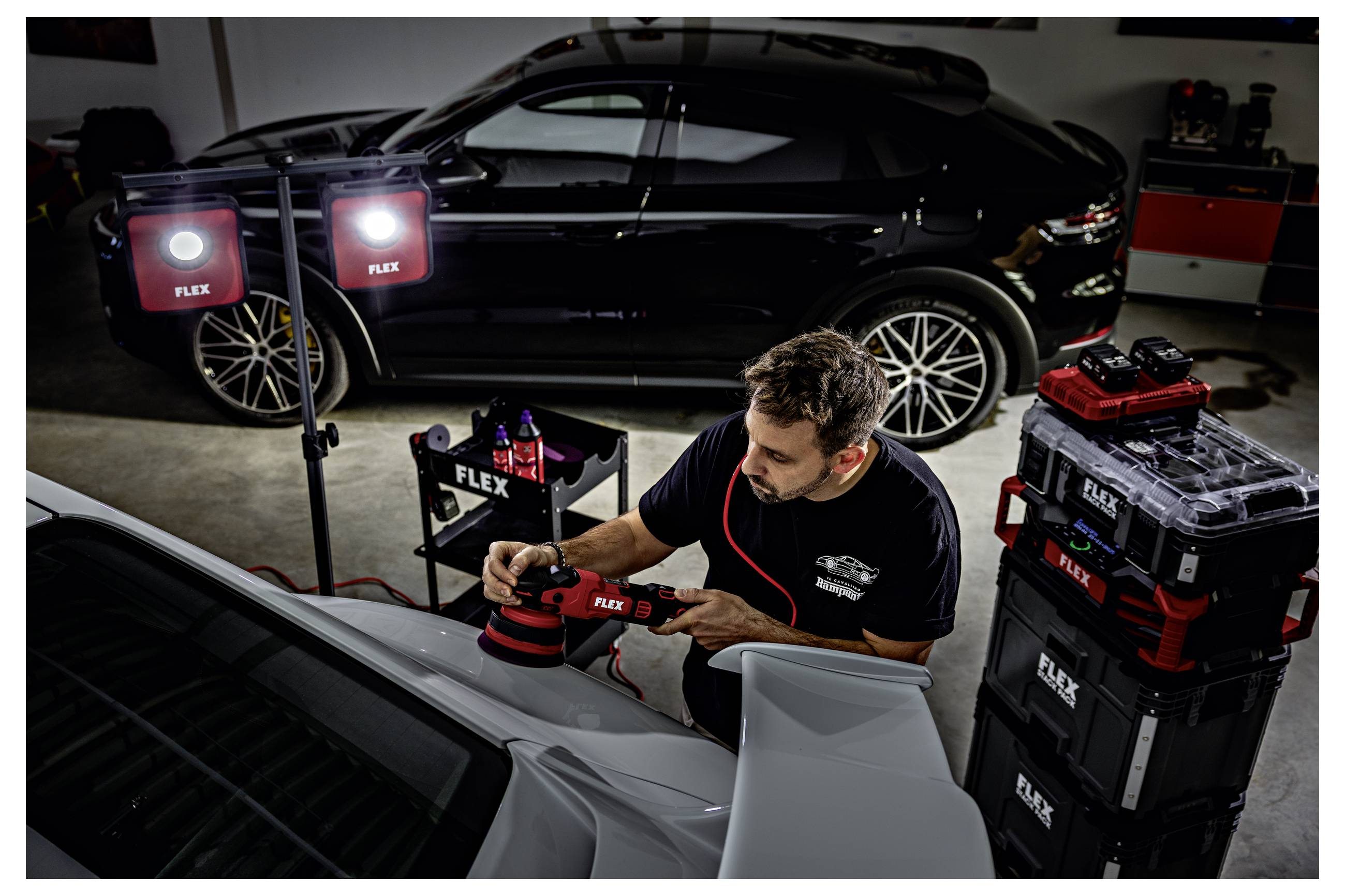 Automechanik leštící bílé auto FLEX leštičkou v garáži vedle černého SUV a při jasném pracovním osvětlení.