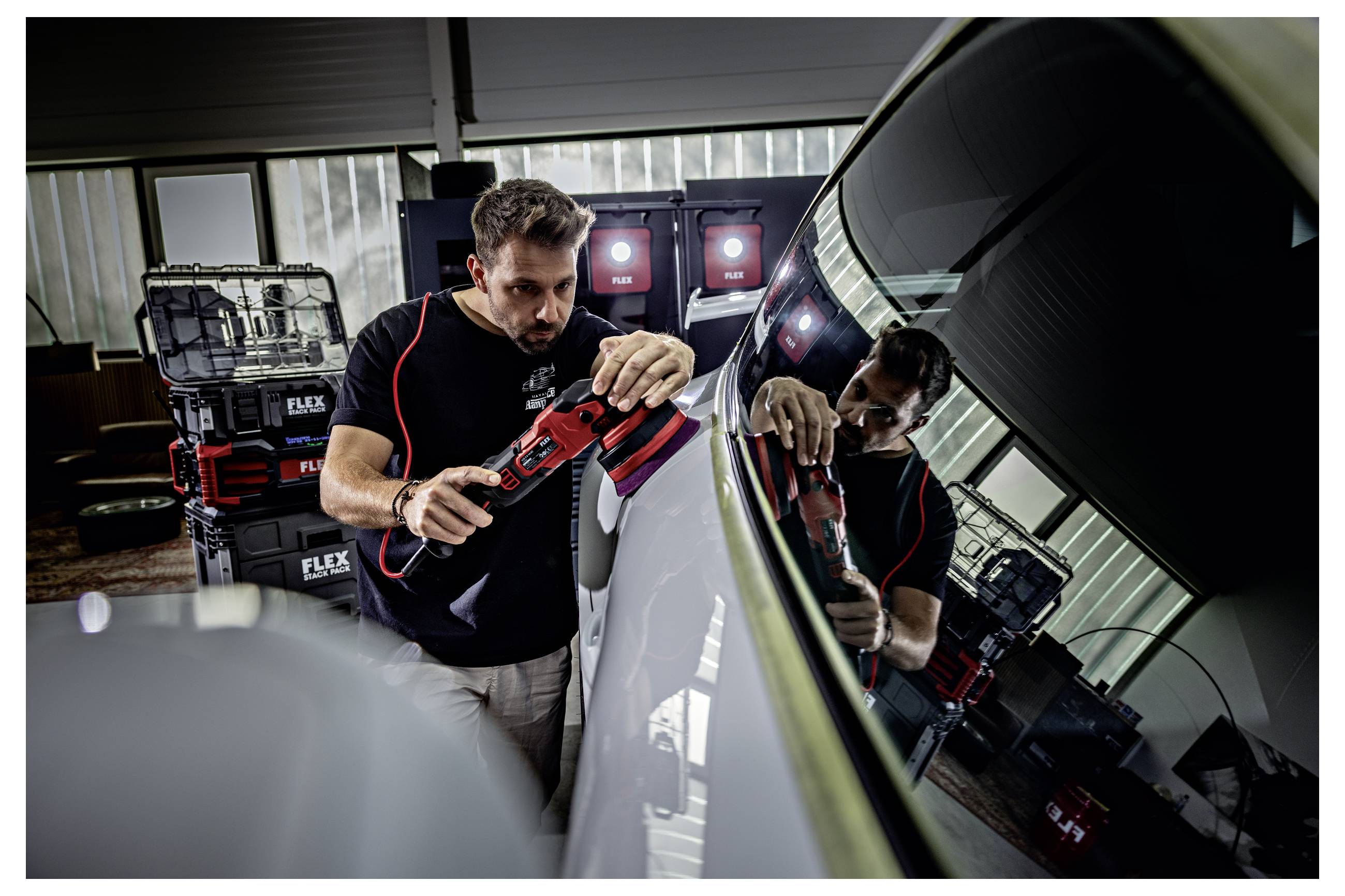 Muž leštící boční panel automobilu červeným elektrickým leštičem v dílně, odrážející se v lesklém nátěru.