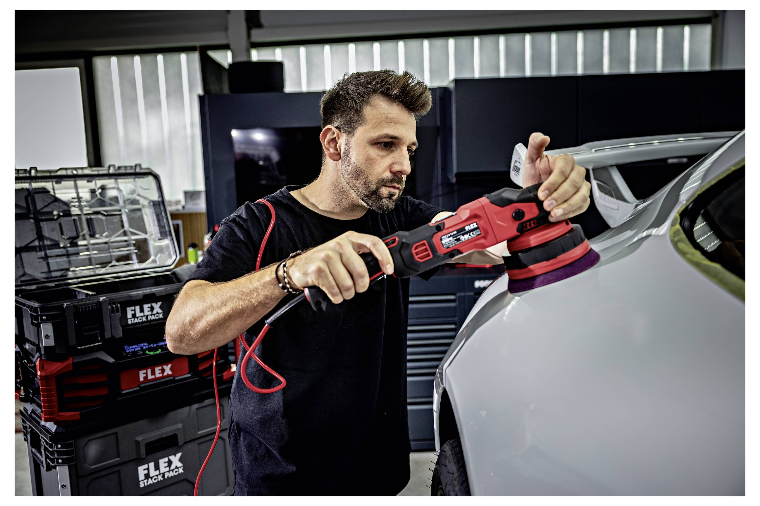 Muž leštící automobilový panel pomocí červeného elektického buferu v autodílně, se skládanými pouzdry nástrojů FLEX v pozadí.