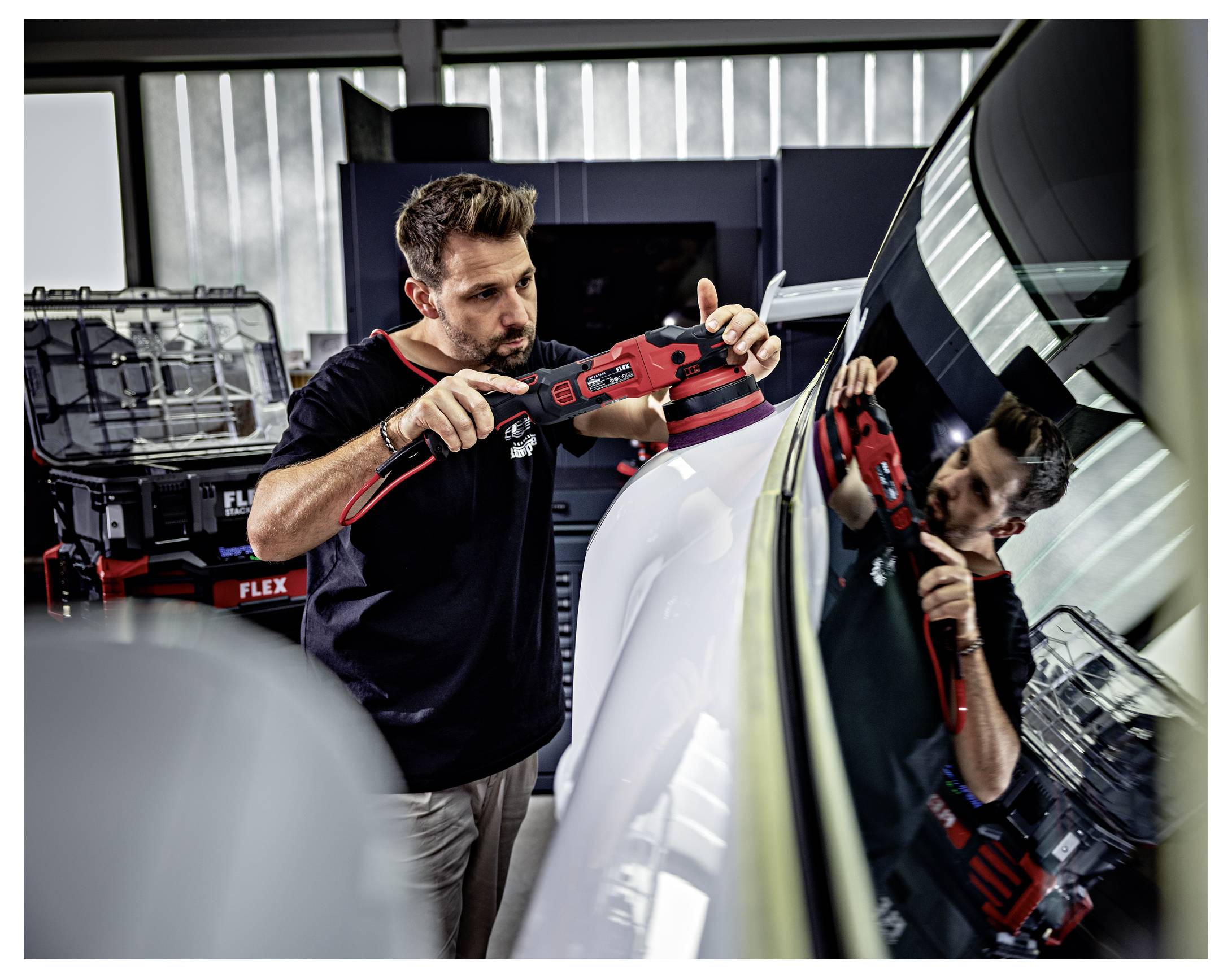 Technik detailingu leštící kapotu automobilu červeným elektrickým leštidlem v dílně, přičemž je vidět nástroj a jeho odraz ve větrném skle.