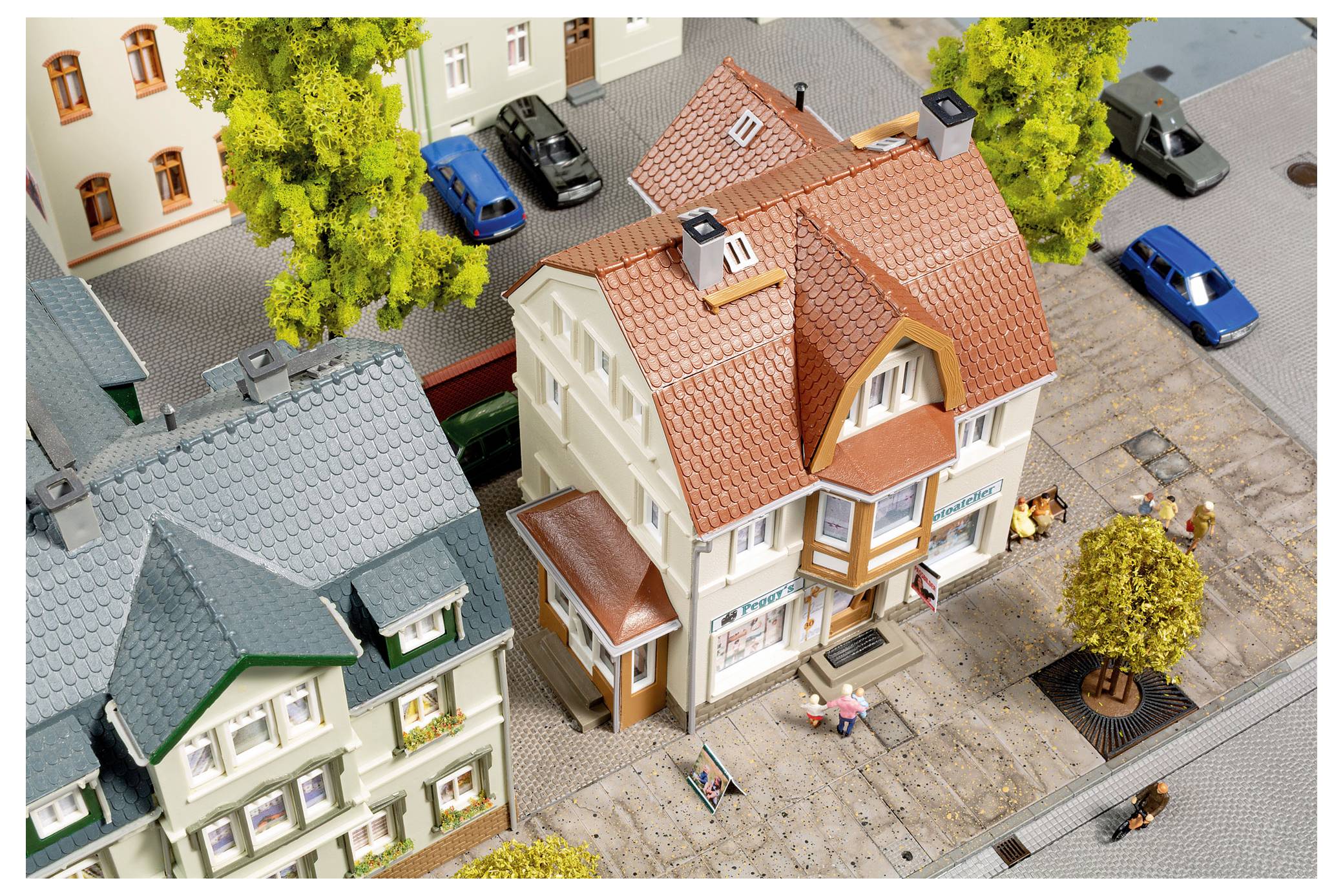 Miniaturní městská scéna s modelovými domy, zaparkovanými auty, stromy a malými figurkami na chodnících, pohled shora.