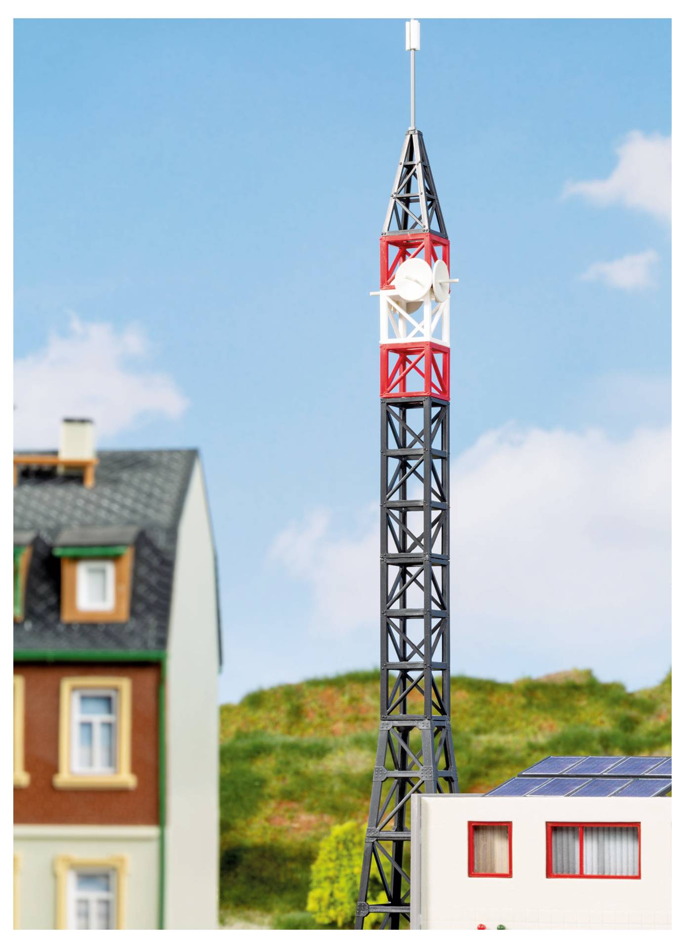 Modelová věž s červenobílým vrcholem stojí vedle miniaturních domků a střechy se solárními panely pod modrým nebem, což naznačuje měřítko modelu osady nebo telekomunikační stanice.