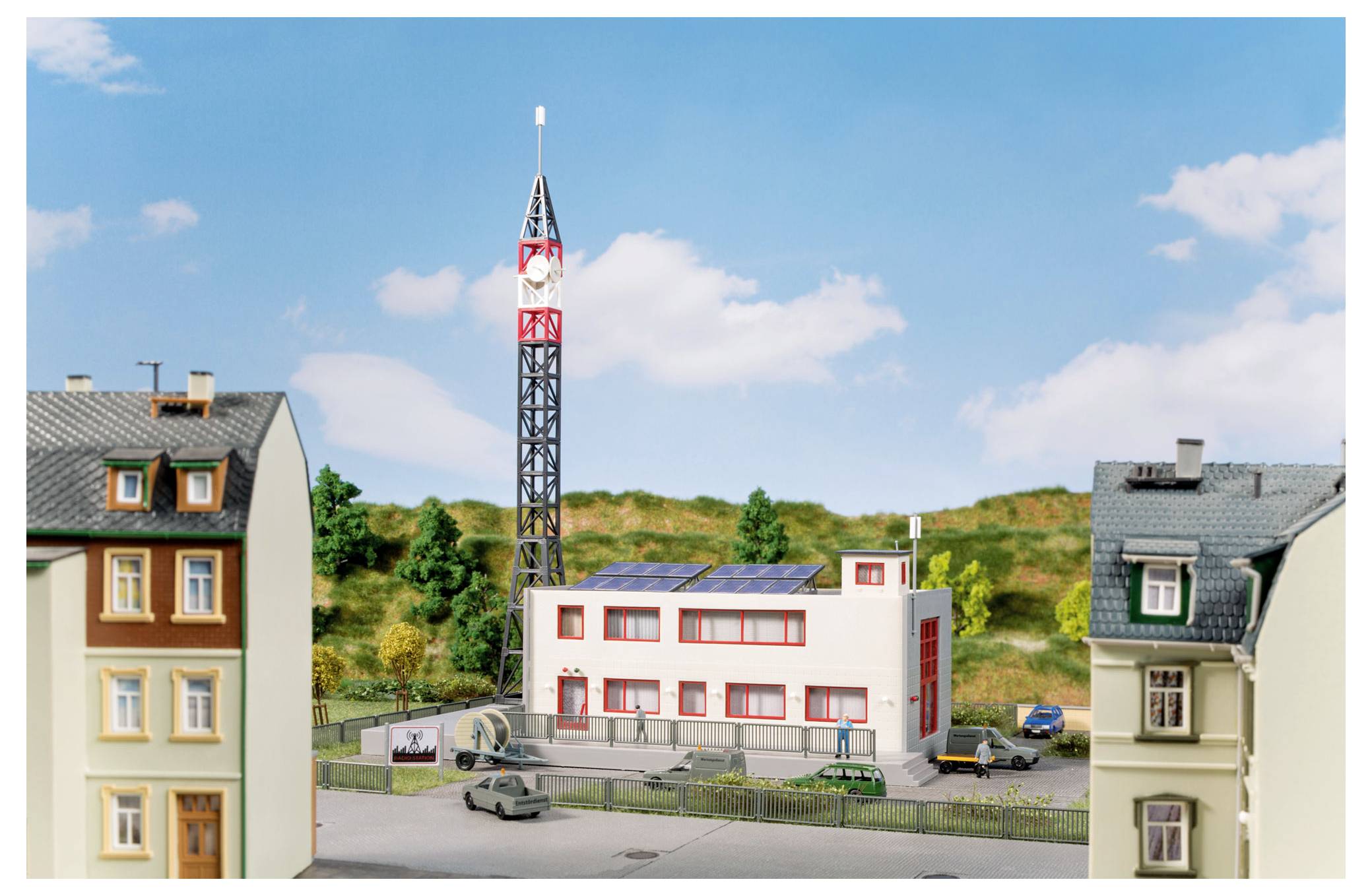 Model malého městského bloku s vysokou radioovou věží, centrální moderní budovou se solárními panely a obklopujícími domy pod modrou oblohou.