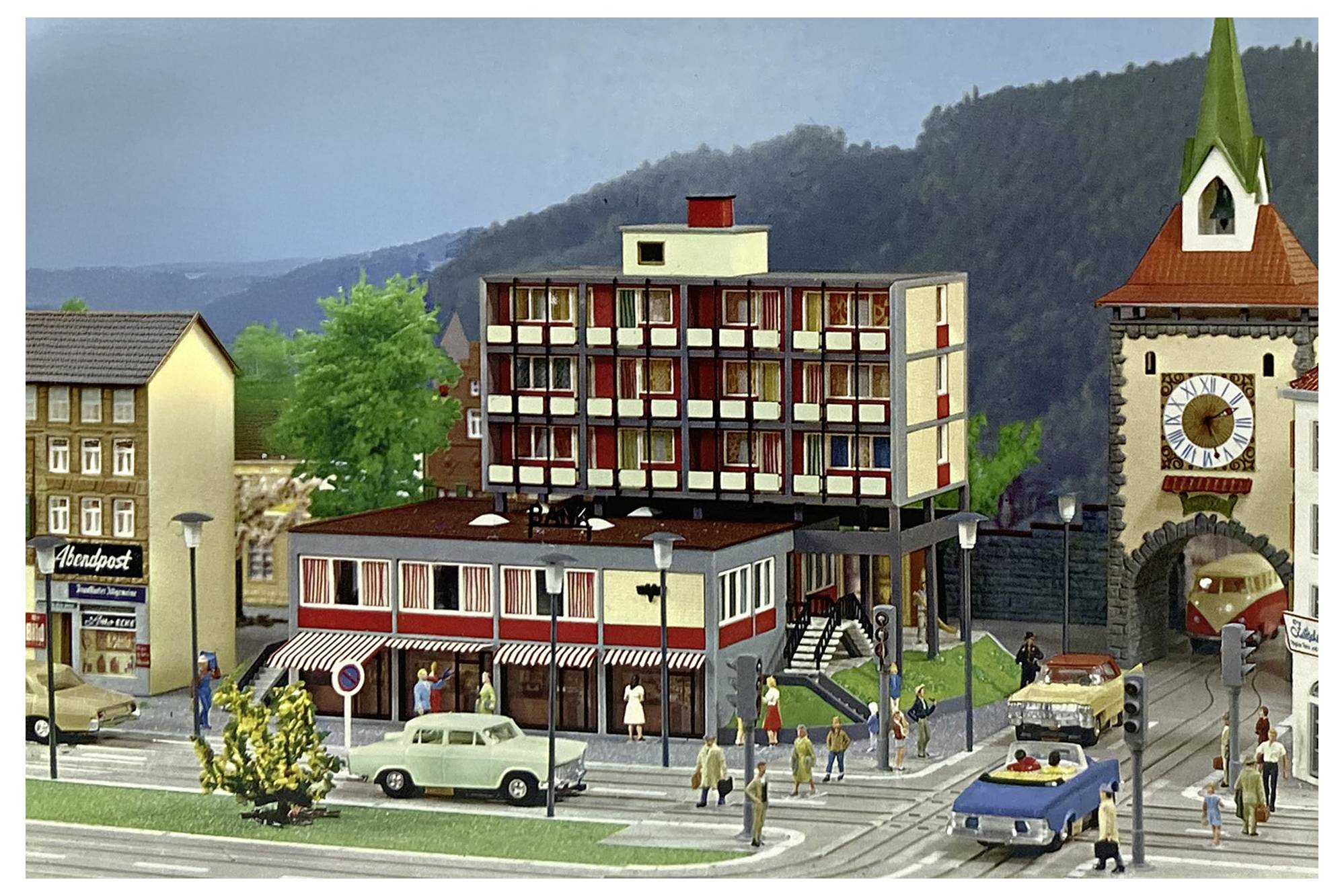 Modelová scéna městské ulice s chodci, automobily, centrální budovou hotelu a věží se hodinami u pozadí hor.