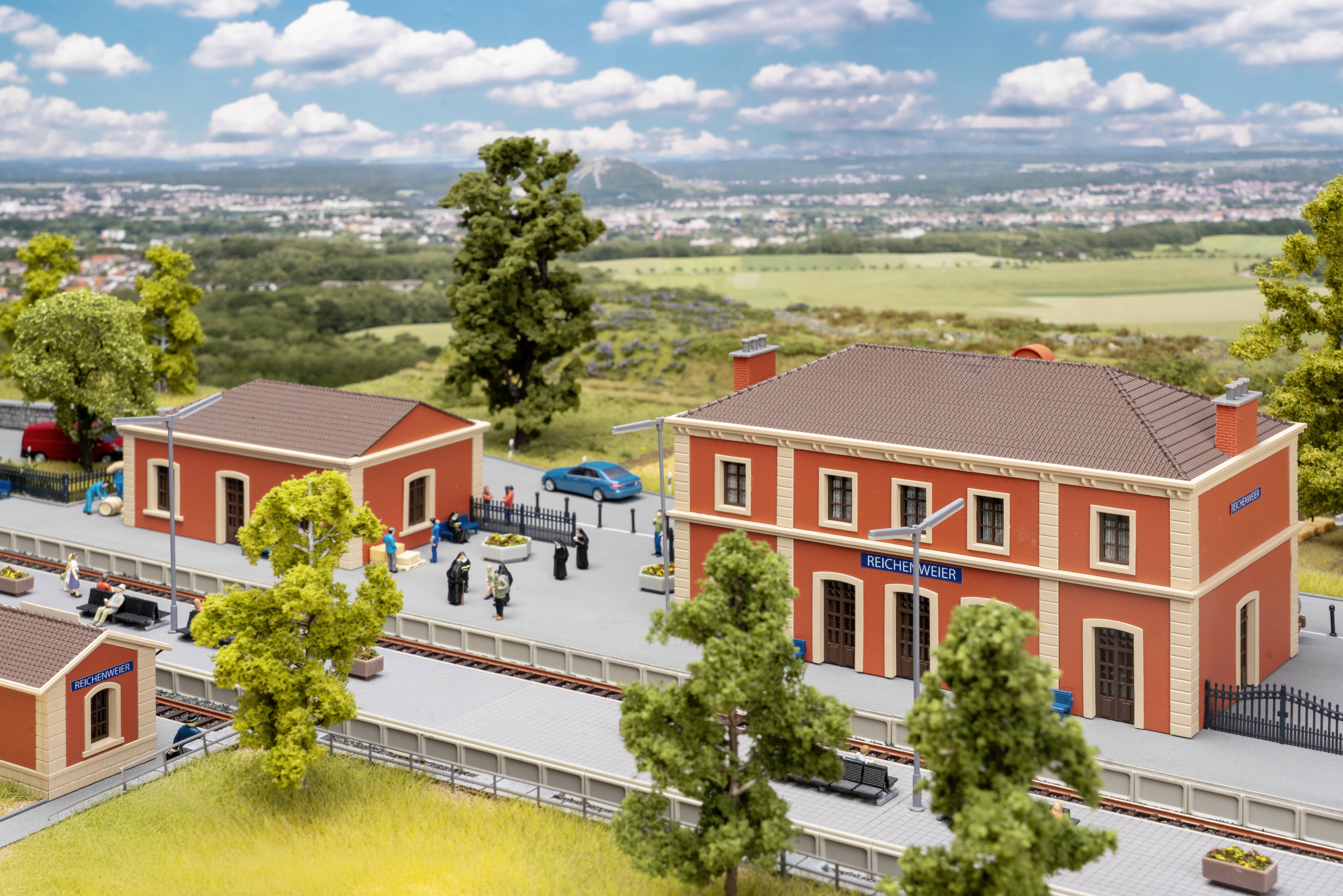 Miniaturní model železniční stanice Beechy View s oranžovými budovami, nástupištěm, lidmi, auty a kolejnicemi v rurální krajině.