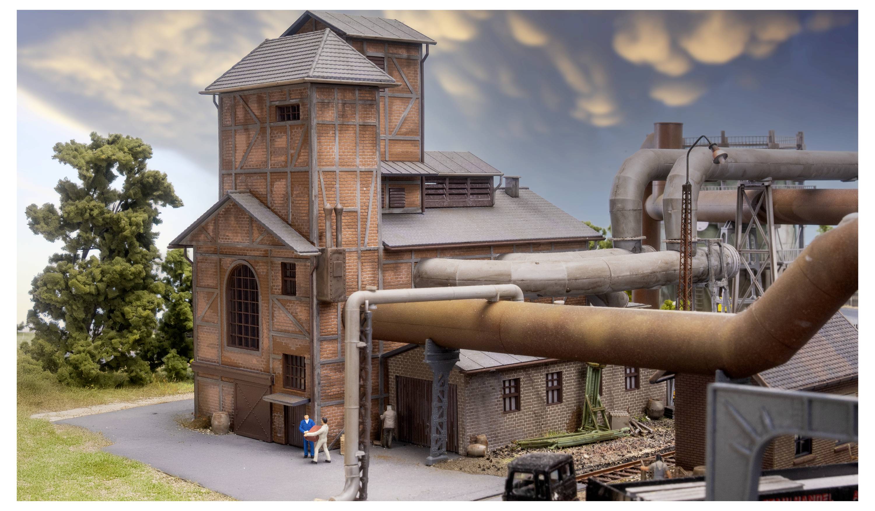 Podrobný model průmyslového závodu s rezavými trubkami, cihlářskými věžemi a malými figurkami vedle budovy.