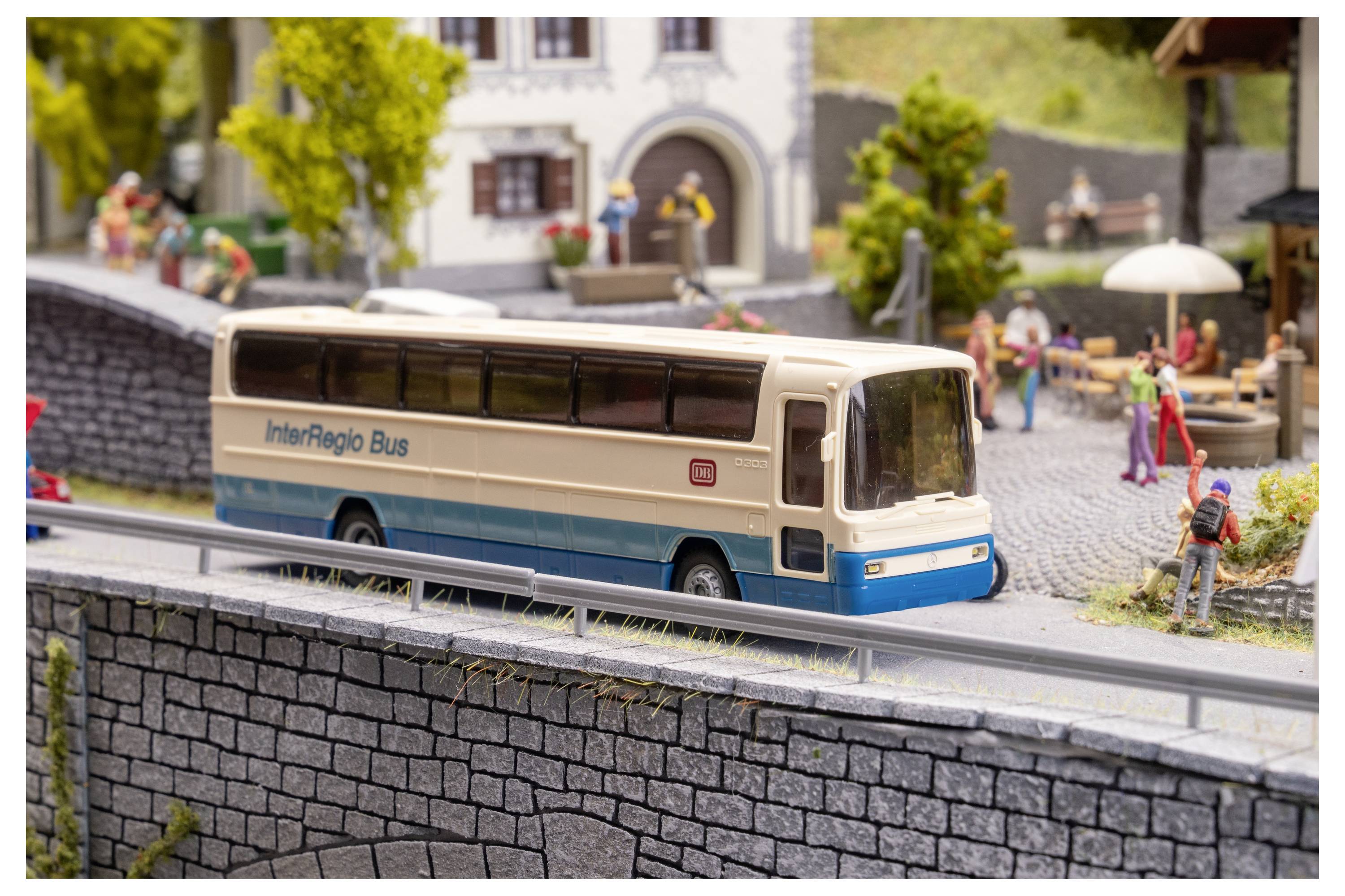 Model meziměstského autobusu na miniaturní vesniční ulici, projíždějící kolem chodců a domů v detailním diorámatu.