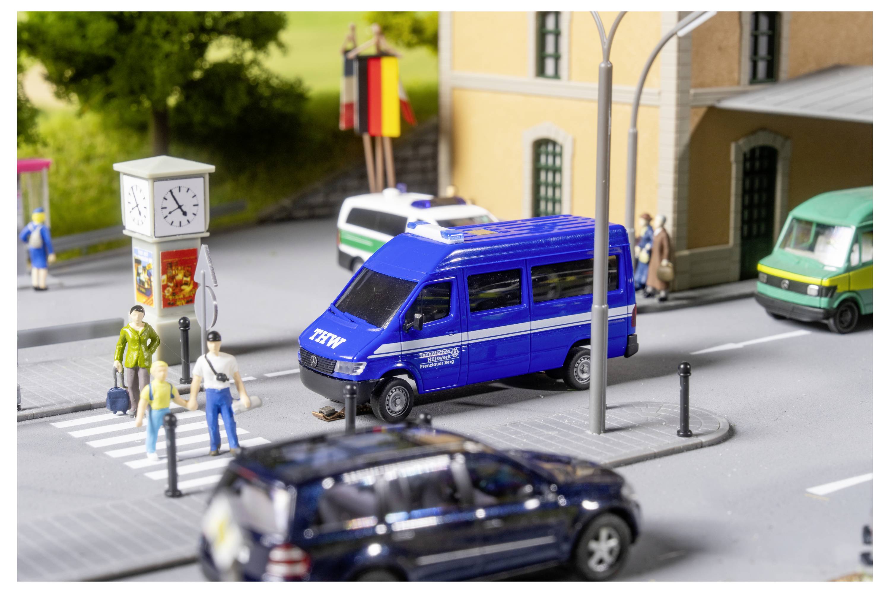 Miniaturní pouliční scéna s modrou policejní dodávkou zastavující se na přechodu pro chodce poblíž chodců, stánku s hodinami a budov; rozmazané auto projíždí v popředí.