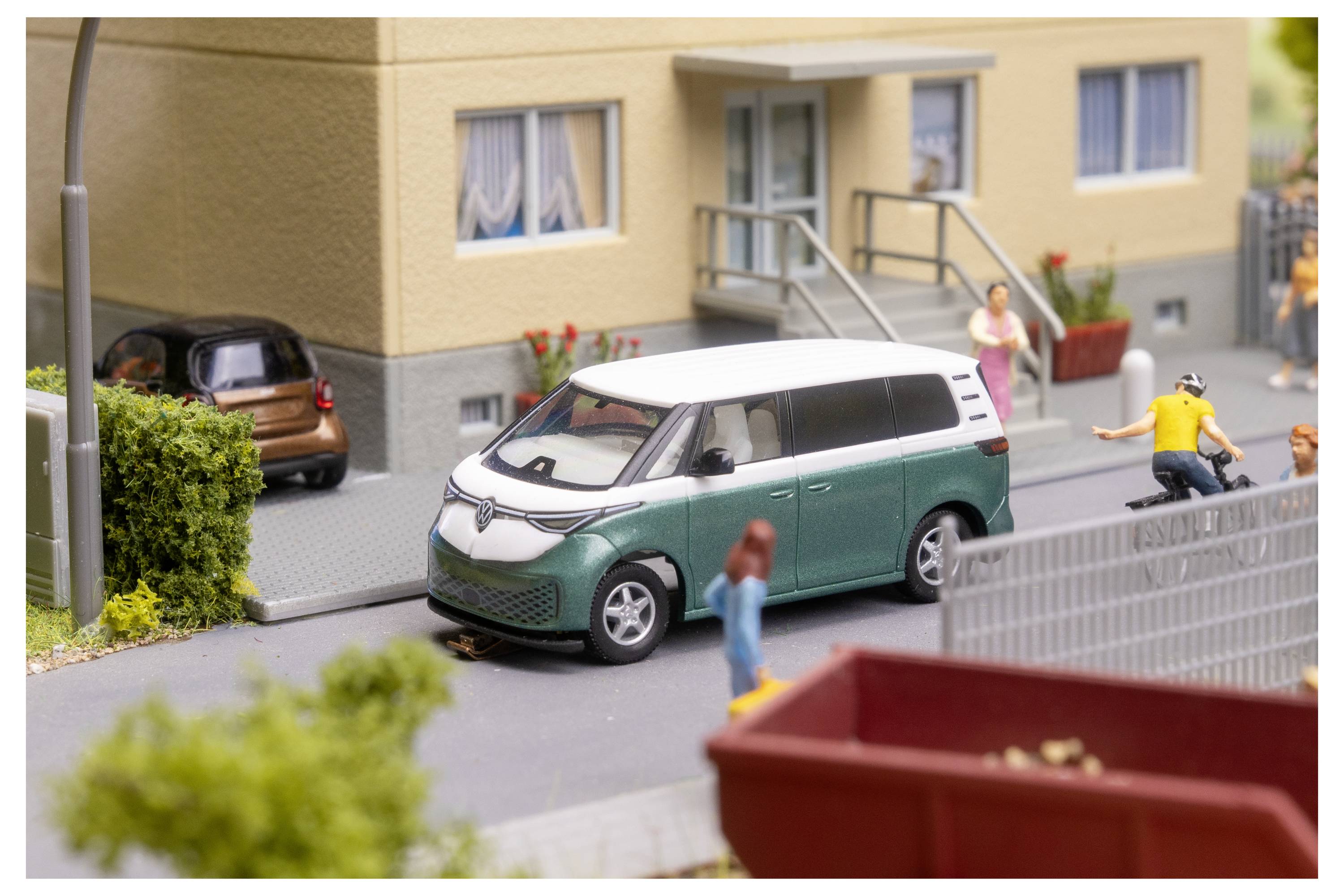 Miniaturní městská scéna s bílým a zeleným dodávkou zaparkovanou u budovy, cyklistou, chodci a zaparkovaným autem v pozadí.