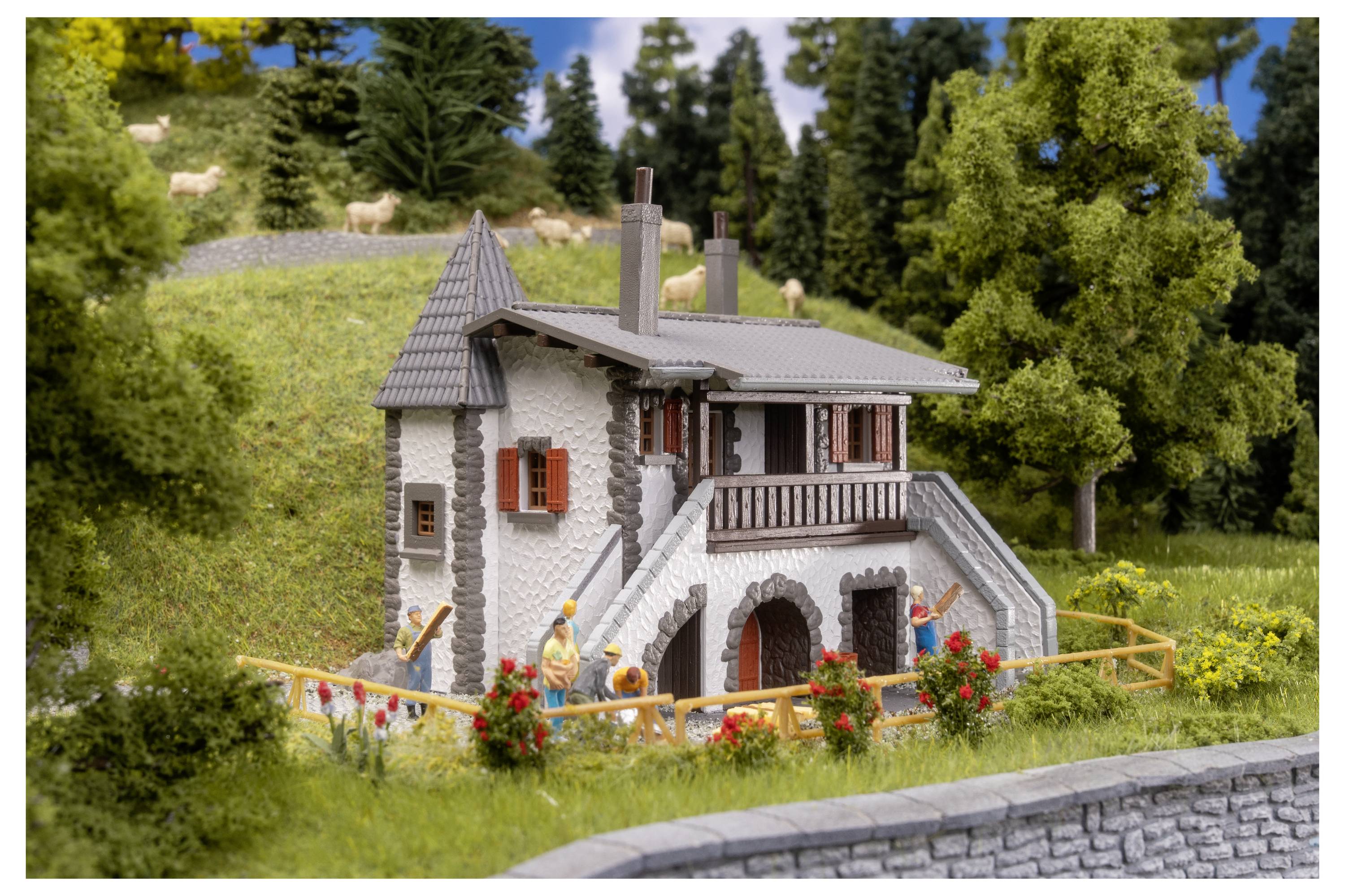 Miniaturní model kamenného domu se schodištěm a balkonem, obklopený trávou, květinami, stromy a malými figurkami v upravené venkovní scéně.