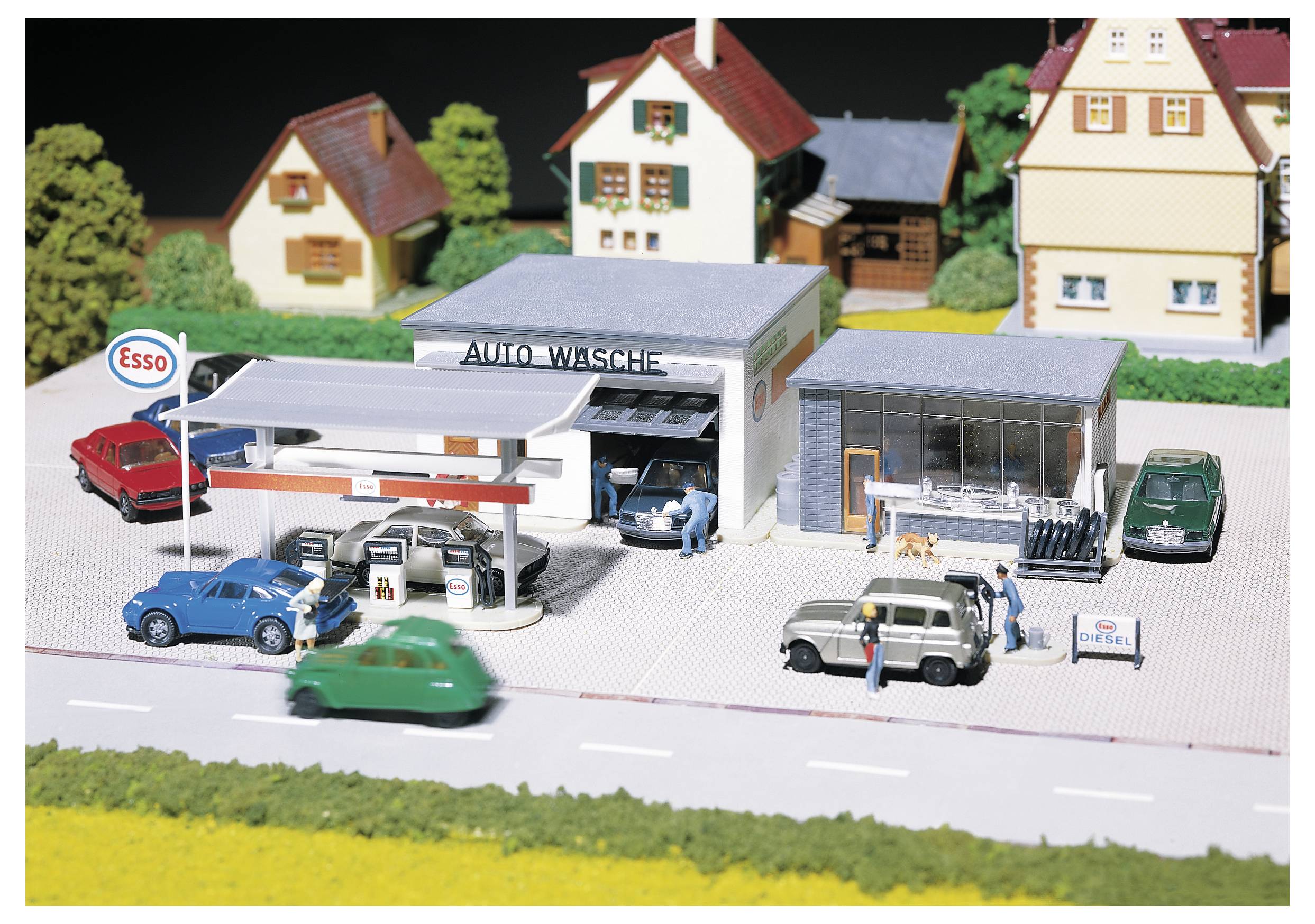 Miniaturní model čerpací stanice a automatické myčky s auty, čerpacími pistolemi a malými figurkami v prostředí vesnice.