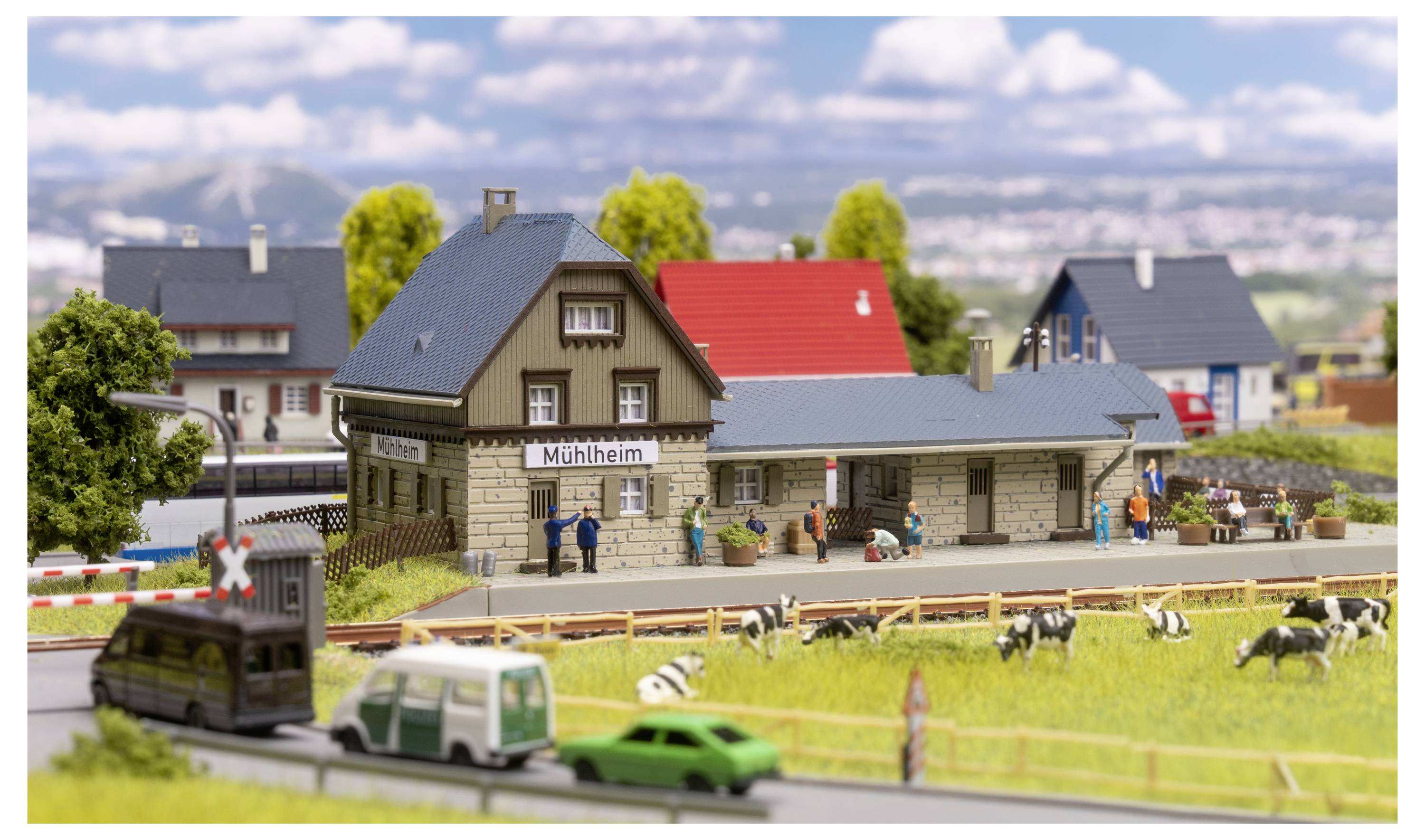 Scéna modelové vlaňské stanice s označením "Mühlheim" s miniaturními budovami, lidmi, auty a kravami na trávnatém poli.