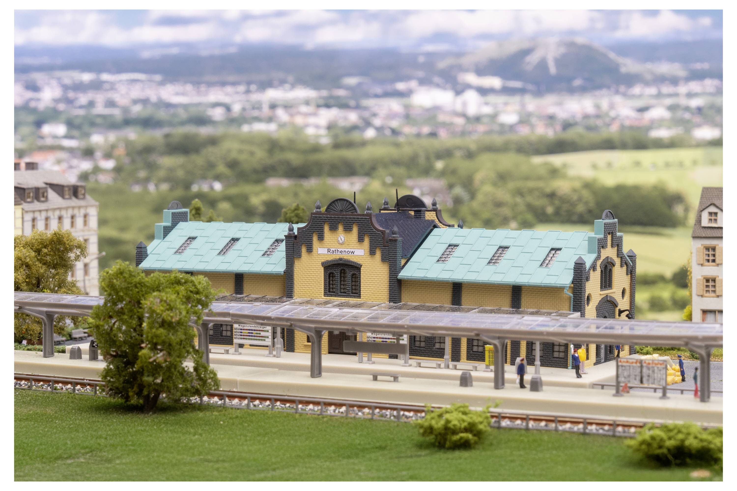 Miniaturní model vlakové stanice s tyrkysovou střechou a nástupištěm, umístěný v modelové vesnici s krajinou a vzdálenými kopci v pozadí.