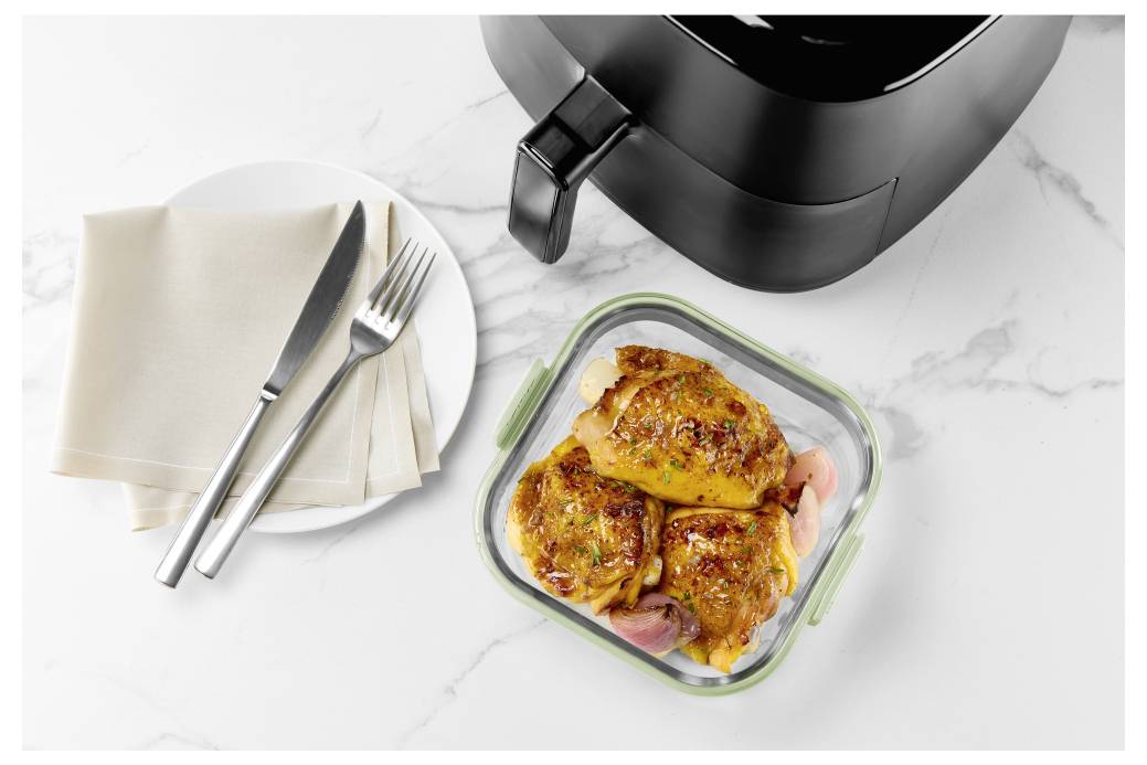 Kontejner na přípravu jídel s připraveným kuřetem v horkovzdušné fritéze, vedle talíře s ubrouskem a příborem na mramorovém kuchyňském pultu.
