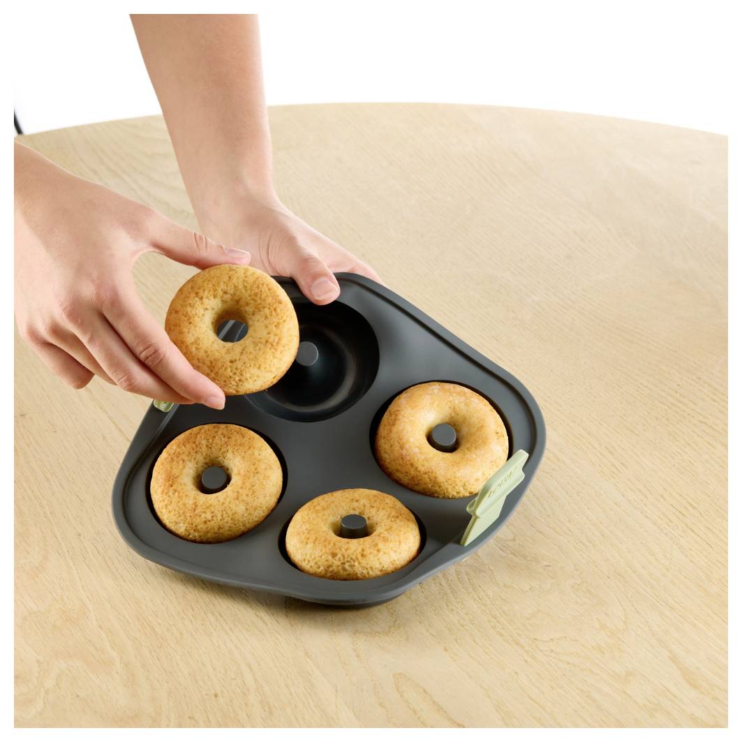 Ruce umisťují pečený donut do černé šestidílné formy na donuchy na dřevěném stole; ve formě jsou již čtyři donuty.