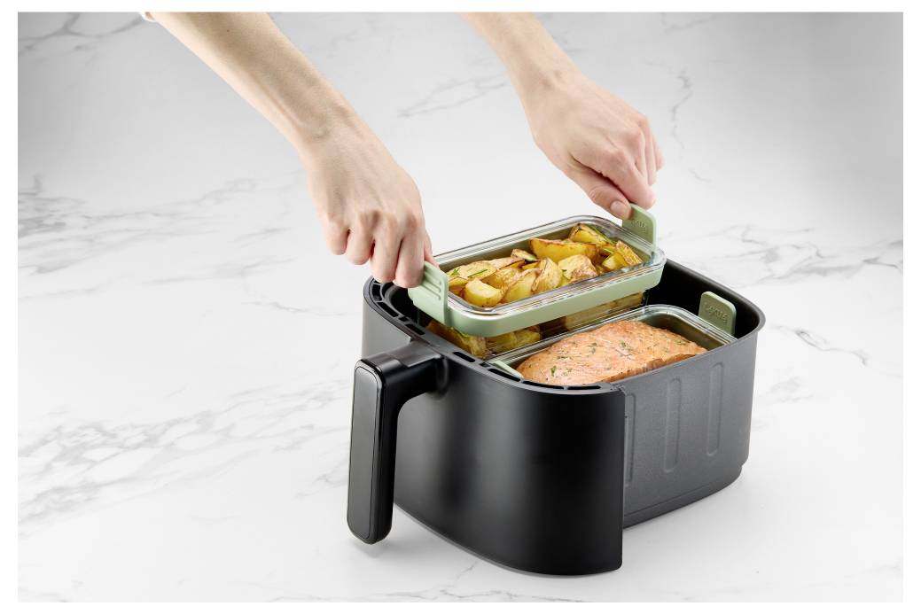 Ruce umisťují skleněnou nádobu na jídlo se pečenými brambory do černého elektrického lunch boxu, který obsahuje vařené losose na bílé pracovní desce.