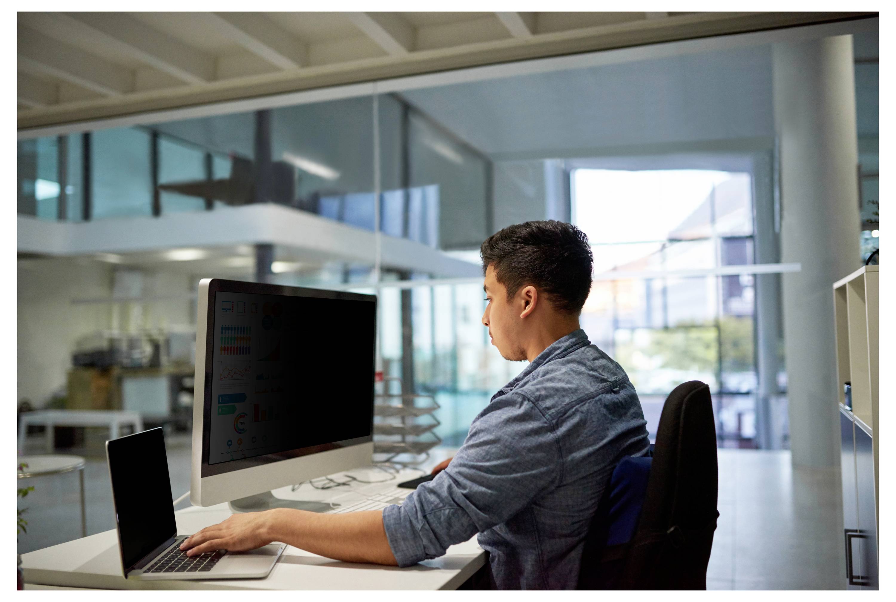 Muž pracující u stolu s laptopem a desktopovým monitorem v moderní kanceláři se skleněnými stěnami.