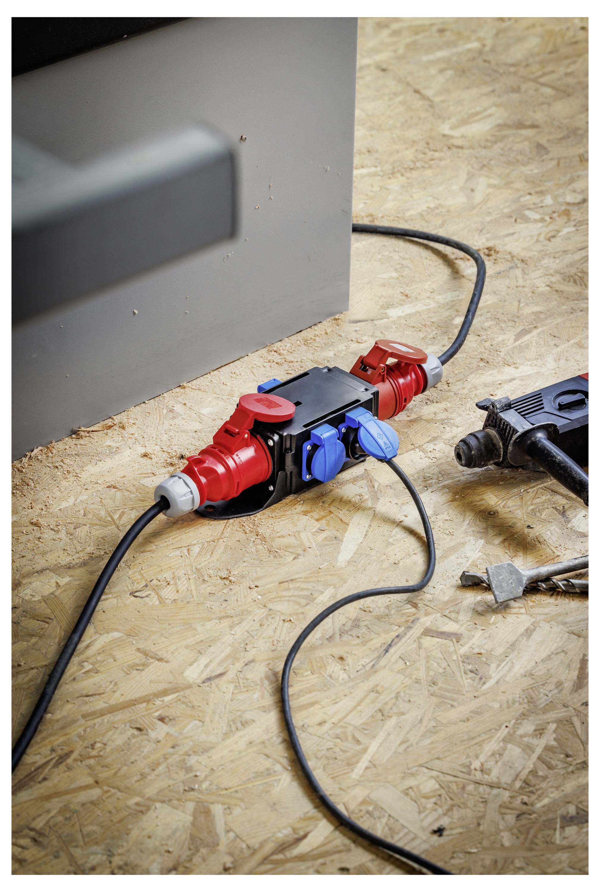 Červený a modrý venkovní elektrický zásuvkový kabel na dřevěné podlaze, s černými kabely připojenými vedle zdi.