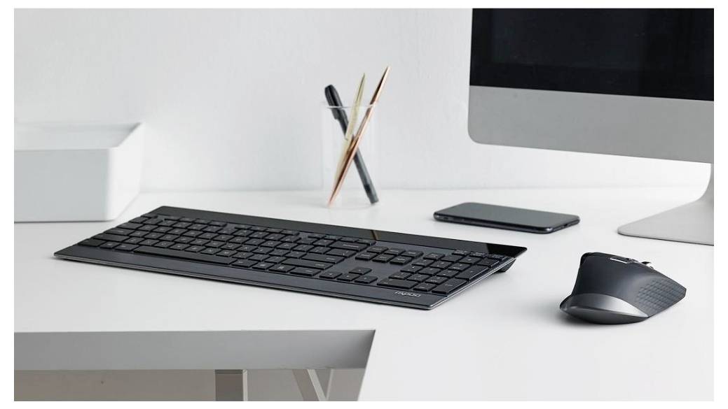 Minimalistické bílé pracovní místo s klávesnicí, myší, chytrým telefonem, kelímkem na tužky a monitorem stolního počítače.