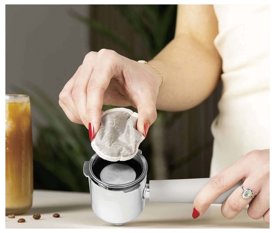 Žena vkládá kávový sáček do kompaktního bílého espressovacího automatu se šedou rukojetí na pracovní desku, nedaleko je chladný nápoj.