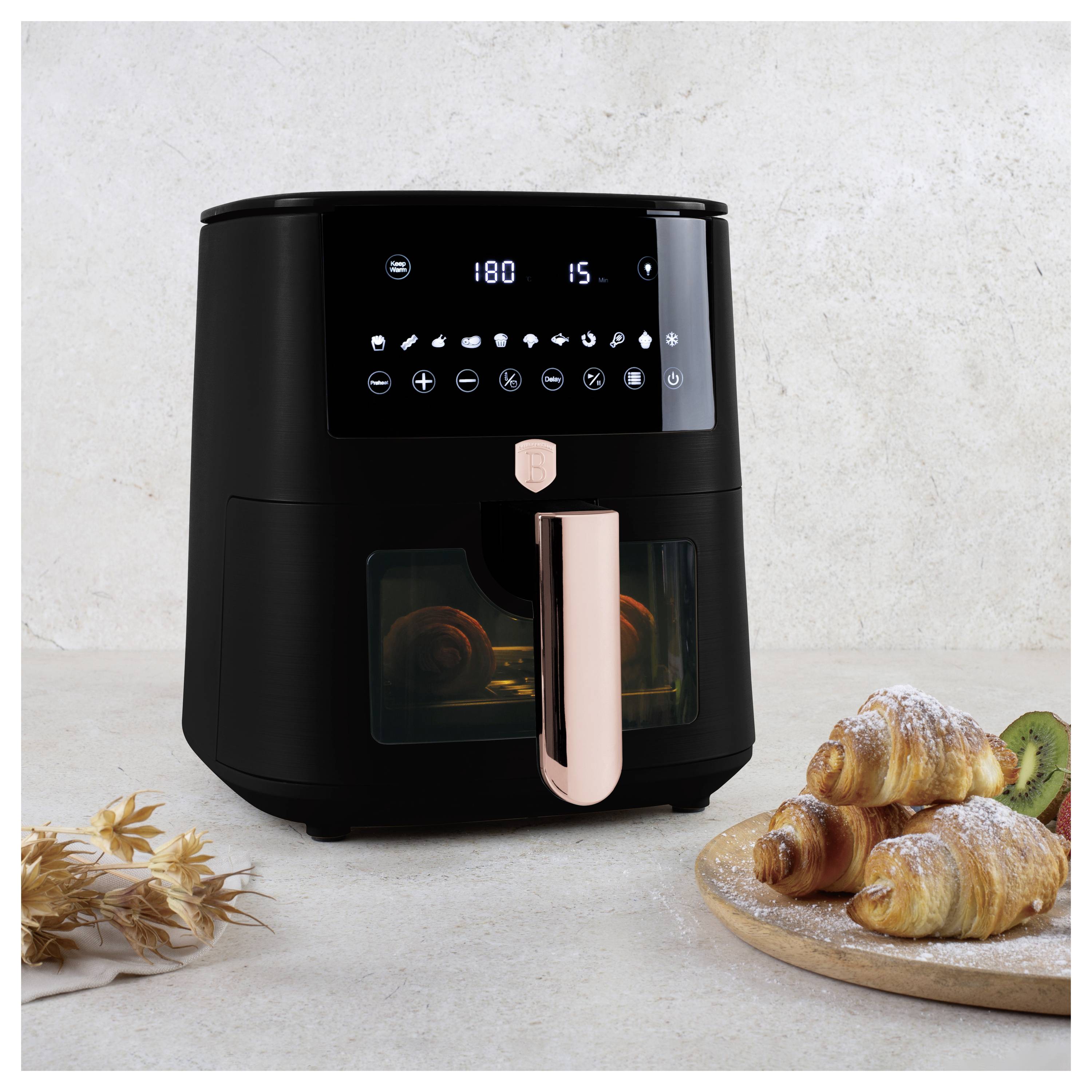 Černá digitální fritéza s růžově zlatým madlem vedle croissantů a kiwi na dřevěné desce.