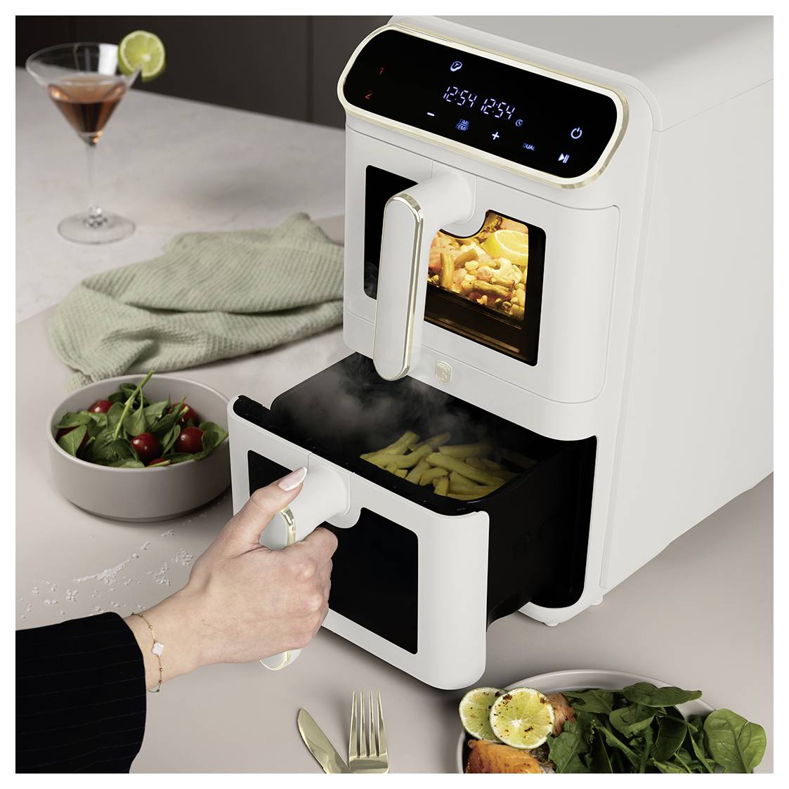 Osoba otevírá dual-zásuvkový horkovzdušný fritér s jídlem vařícím se uvnitř, umístěný na kuchyňský pult s salátkou a koktejlem v blízkosti.