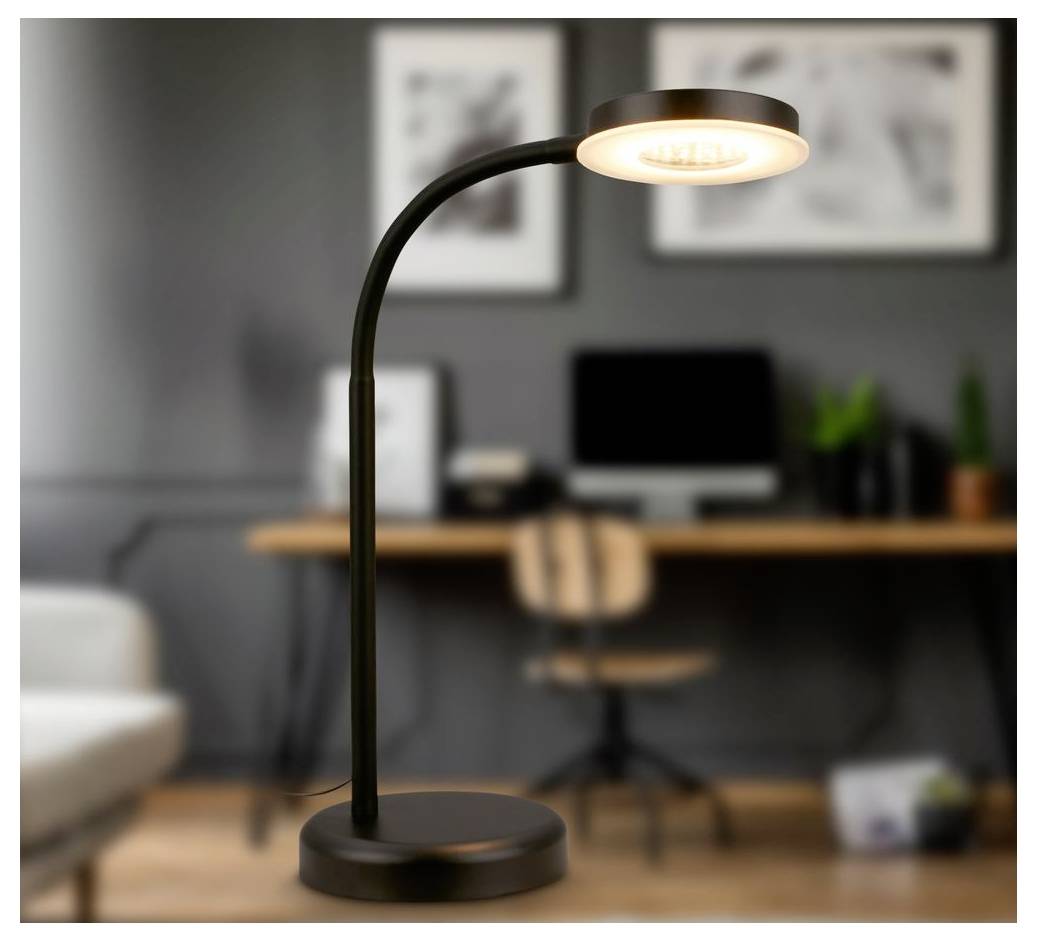Černá stolní lampa s rozsvícenou kulatou hlavicí na kulatém podstavci, zobrazená v rozostřené domácí kanceláři se stolem a židlí v pozadí.