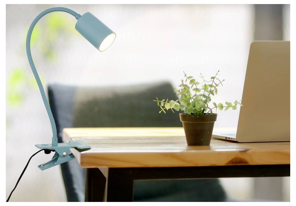 Modrá stolní lampa s klipem osvětlující dřevěný stůl vedle květináče s rostlinou a otevřeného notebooku v domácím pracovním prostoru.