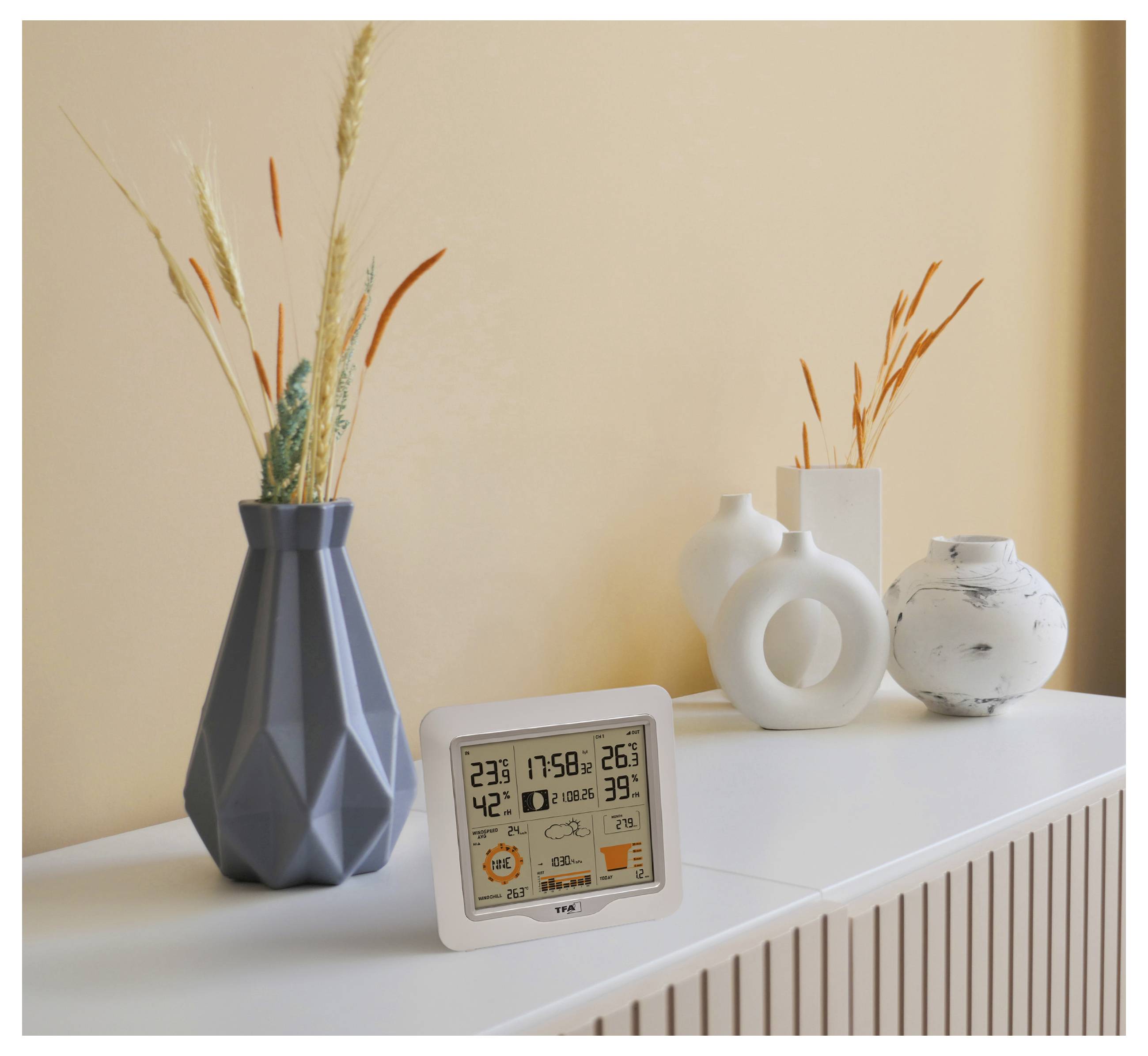 Minimalistické policové uspořádání s digitálním teploměrem/vlhkoměrem, dekorativními vázy a suchými trávami před béžovou stěnou.