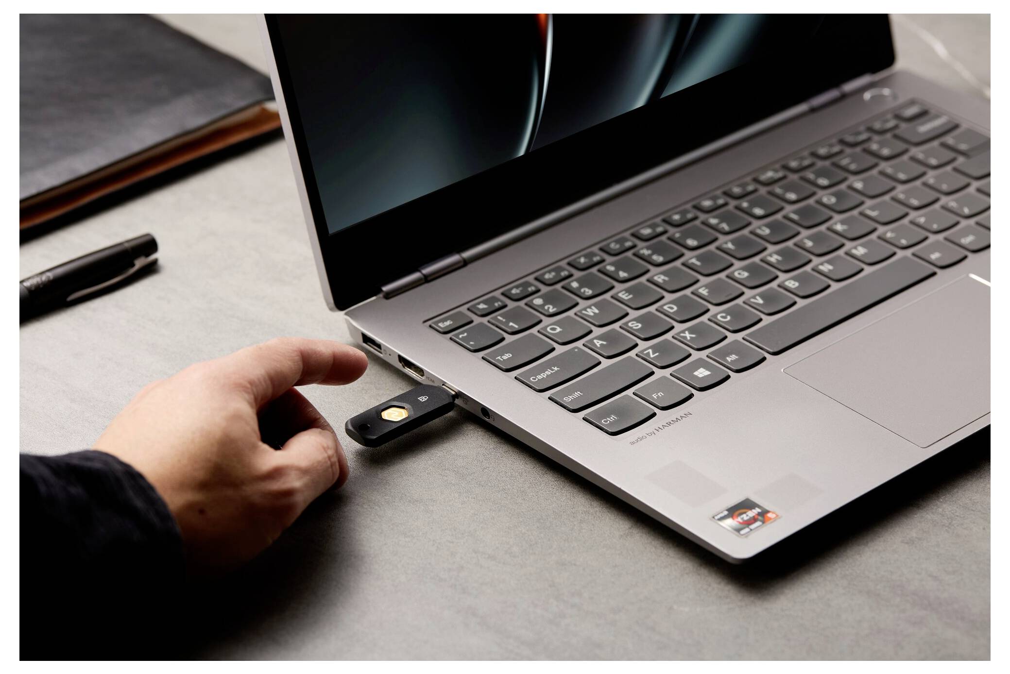 Osoba připojuje USB bezpečnostní klíč do přenosného počítače na stole, což naznačuje двухfaktorové nebo hardwarové ověřování.
