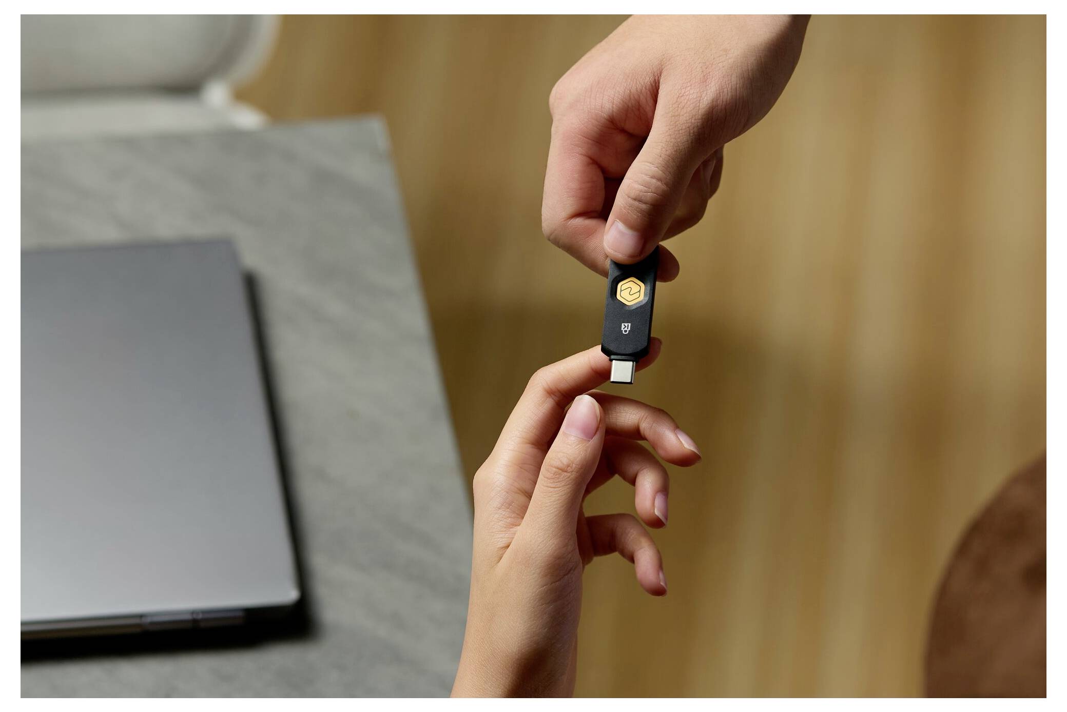 Ruce si vyměňující USB bezpečnostní klíč vedle notebooku, což naznačuje dvoustupňové ověřování nebo bezpečné přihlášení.