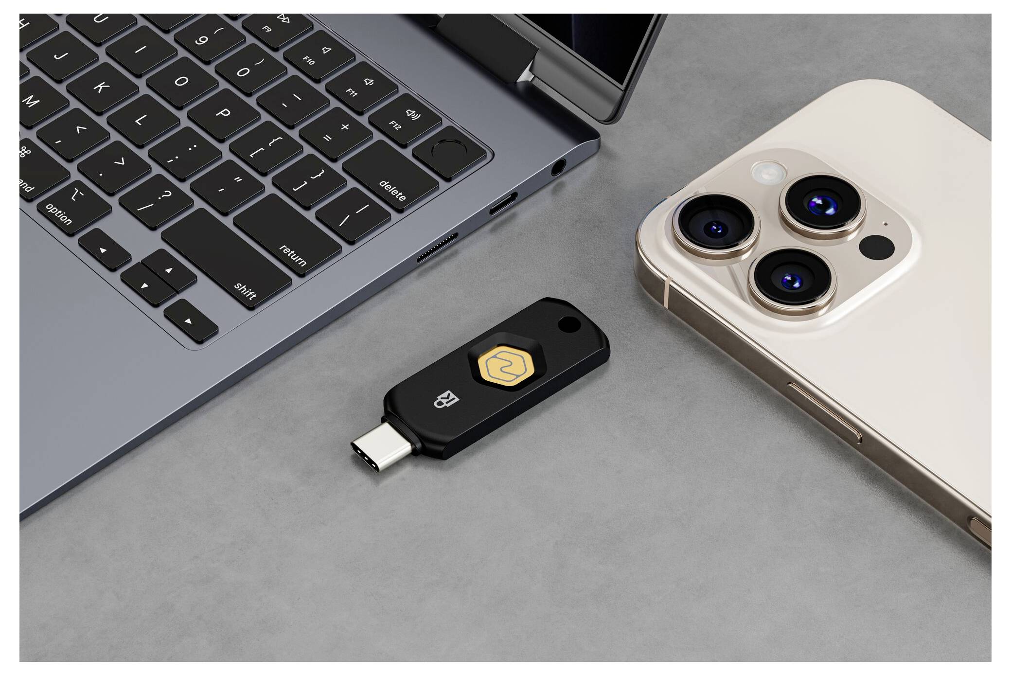 Černý bezpečnostní klíč USB-C mezi notebookem a iPhonem na šedém povrchu, ukazující hardwarový dvoufaktorový ověřovací nástroj pro přihlášení zařízení.
