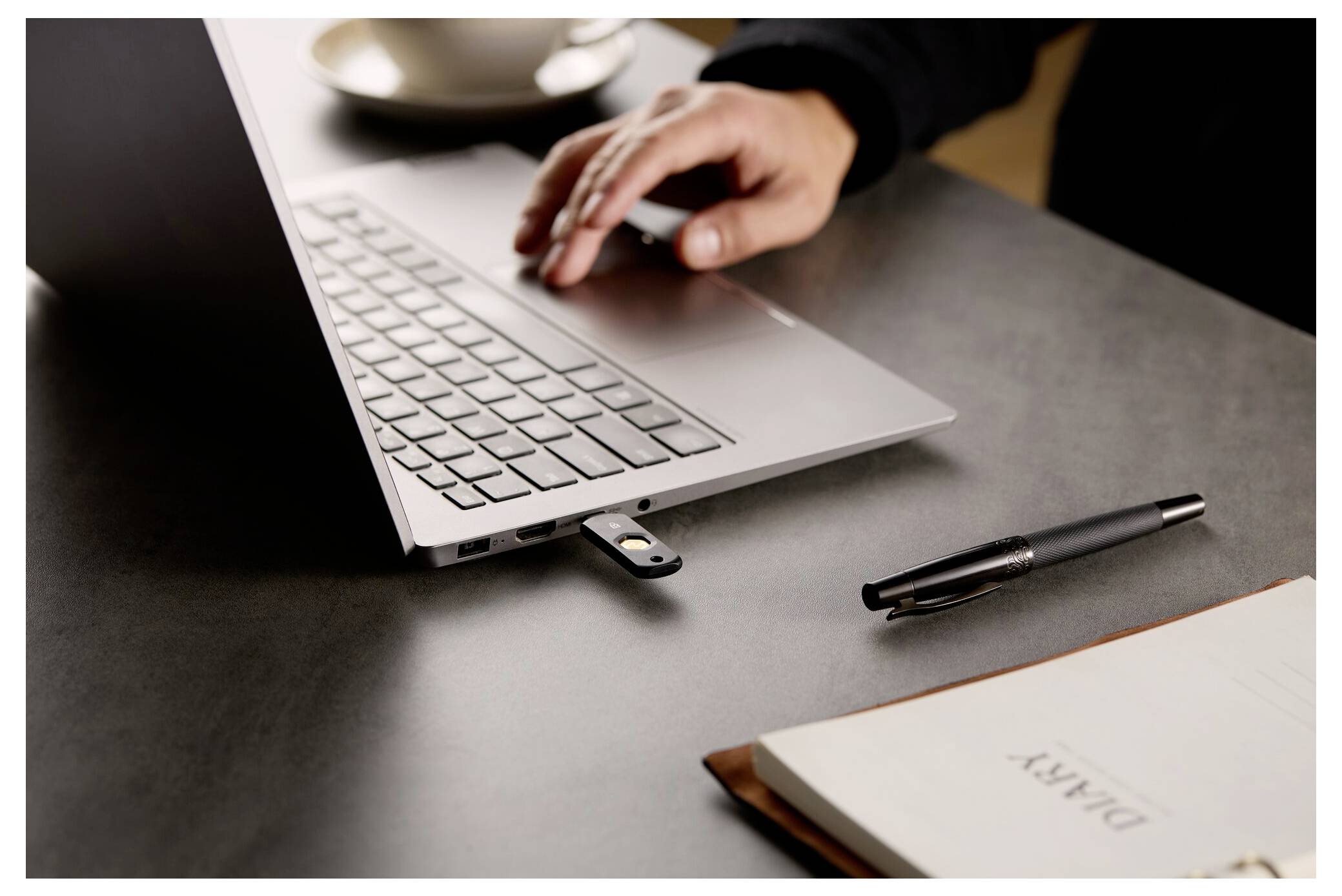 Osoba píšící na notebooku s vloženou USB flash diskou, vedle pera a sešitu s nápisem "DEAR" na stole.