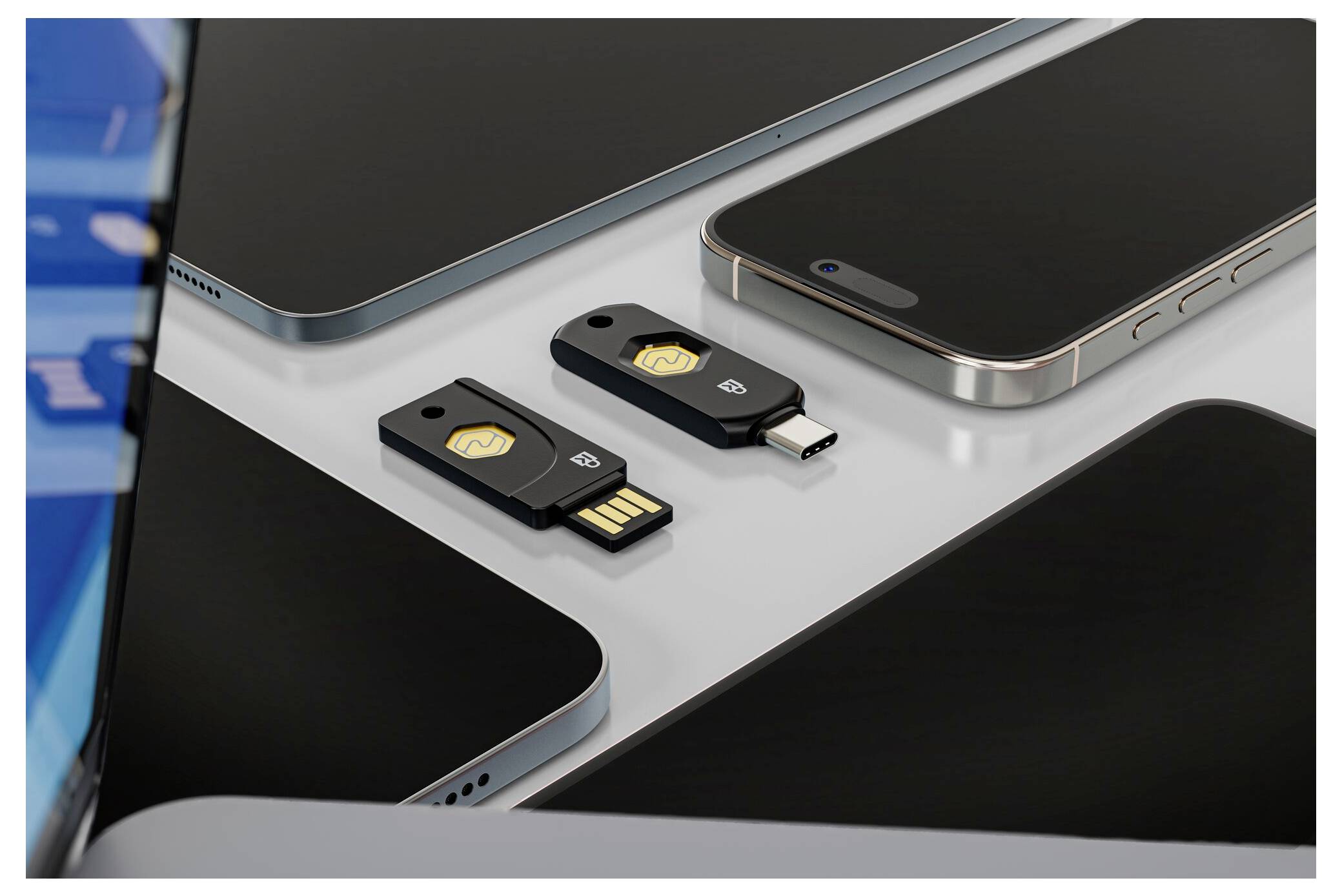 Dva USB bezpečnostní klíče, jeden USB-A a jeden USB-C, umístěné mezi několika chytrými telefony na stole.