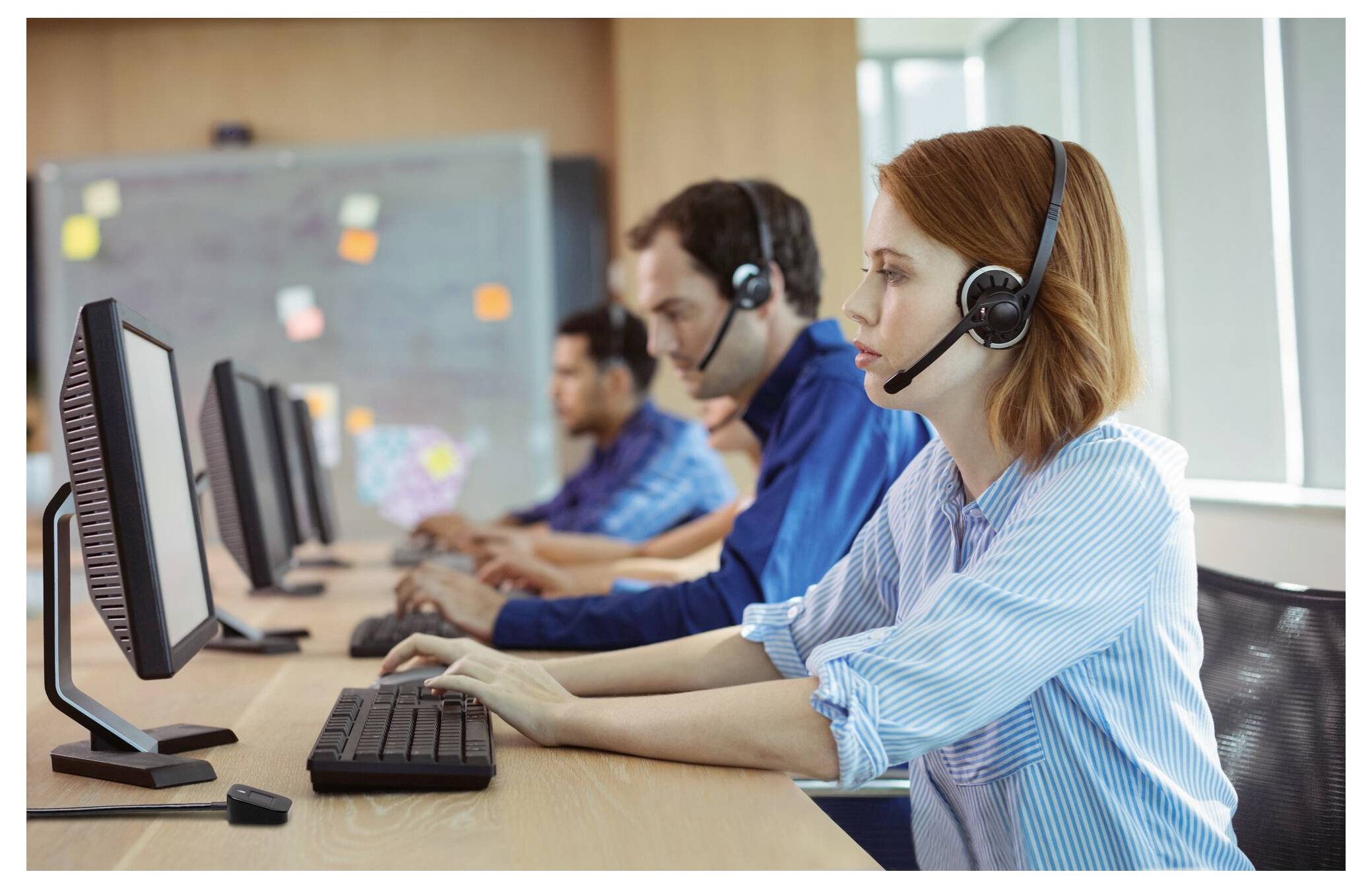 Tři agenti zákaznického servisu v sluchátkách pracují u stolních počítačů v call centru.