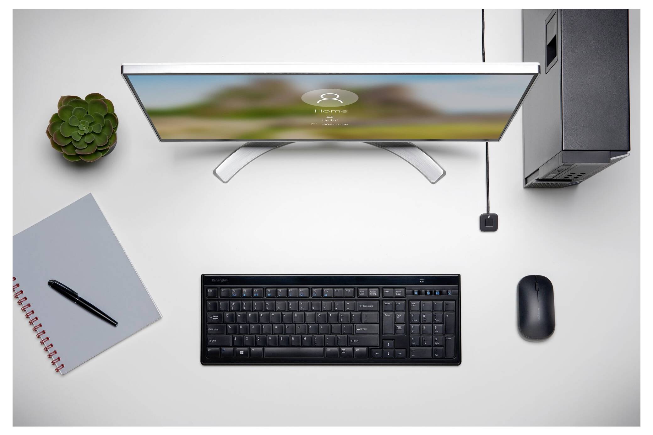 Pohled z vrchu na minimalistický stůl s monitorem, klávesnicí, myší, notebookem a perem, počítačovou věží, elektrickou zásuvkou a květinou v květináči.