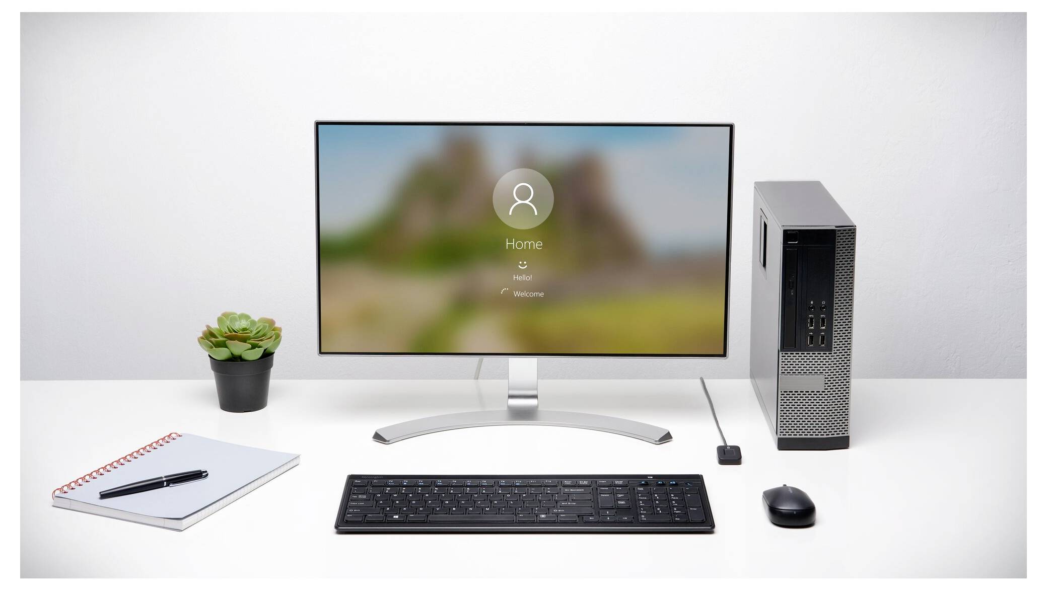 Nastavení stolního počítače s monitorem zobrazujícím přihlašovací obrazovku, klávesnicí, myší, malou rostlinou, notebookem a věží PC na bílém stole.