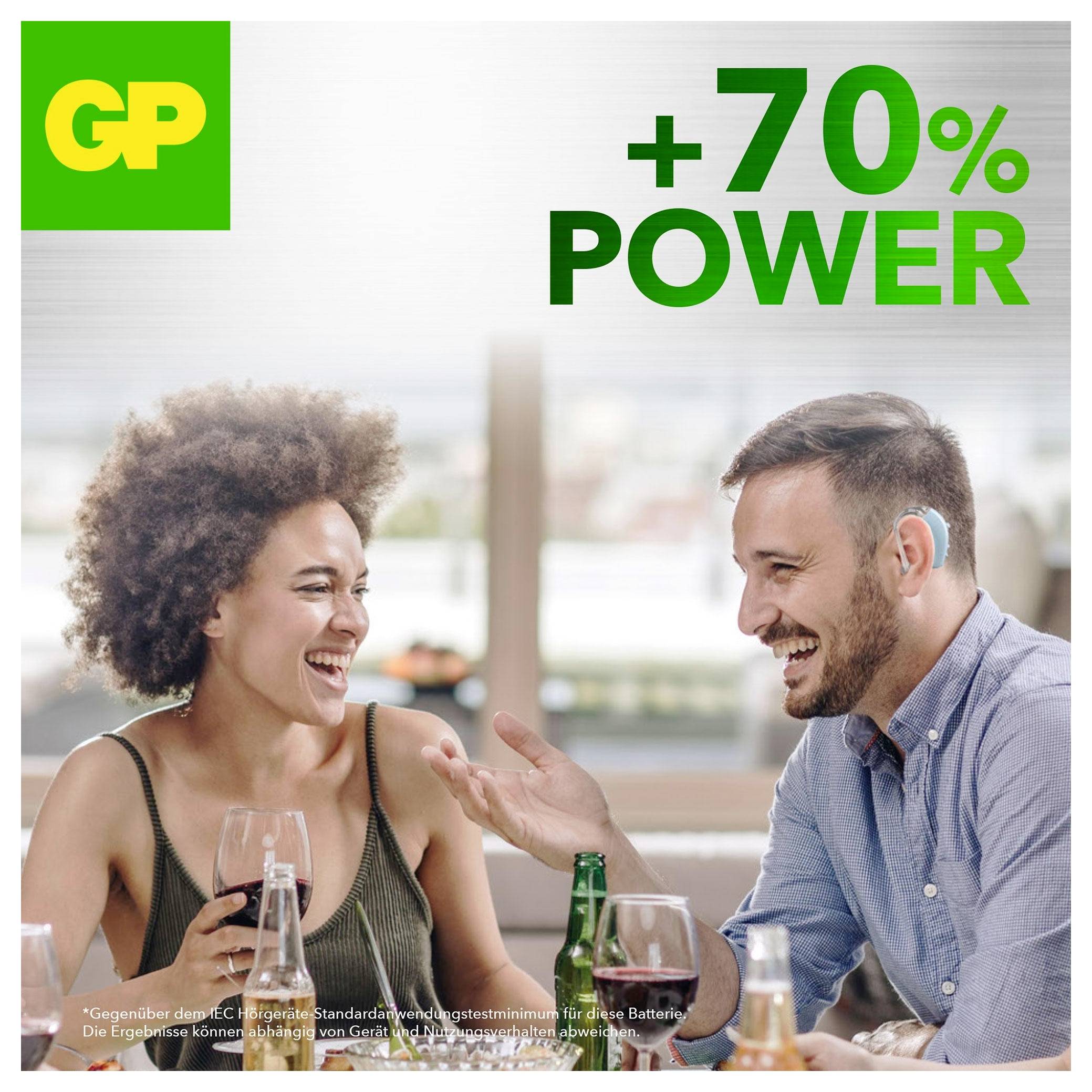Dva lidé se usměvají a mluví u stolu s GP brandingem a textem „+70% POWER" propagující zvýšený výkon baterie.