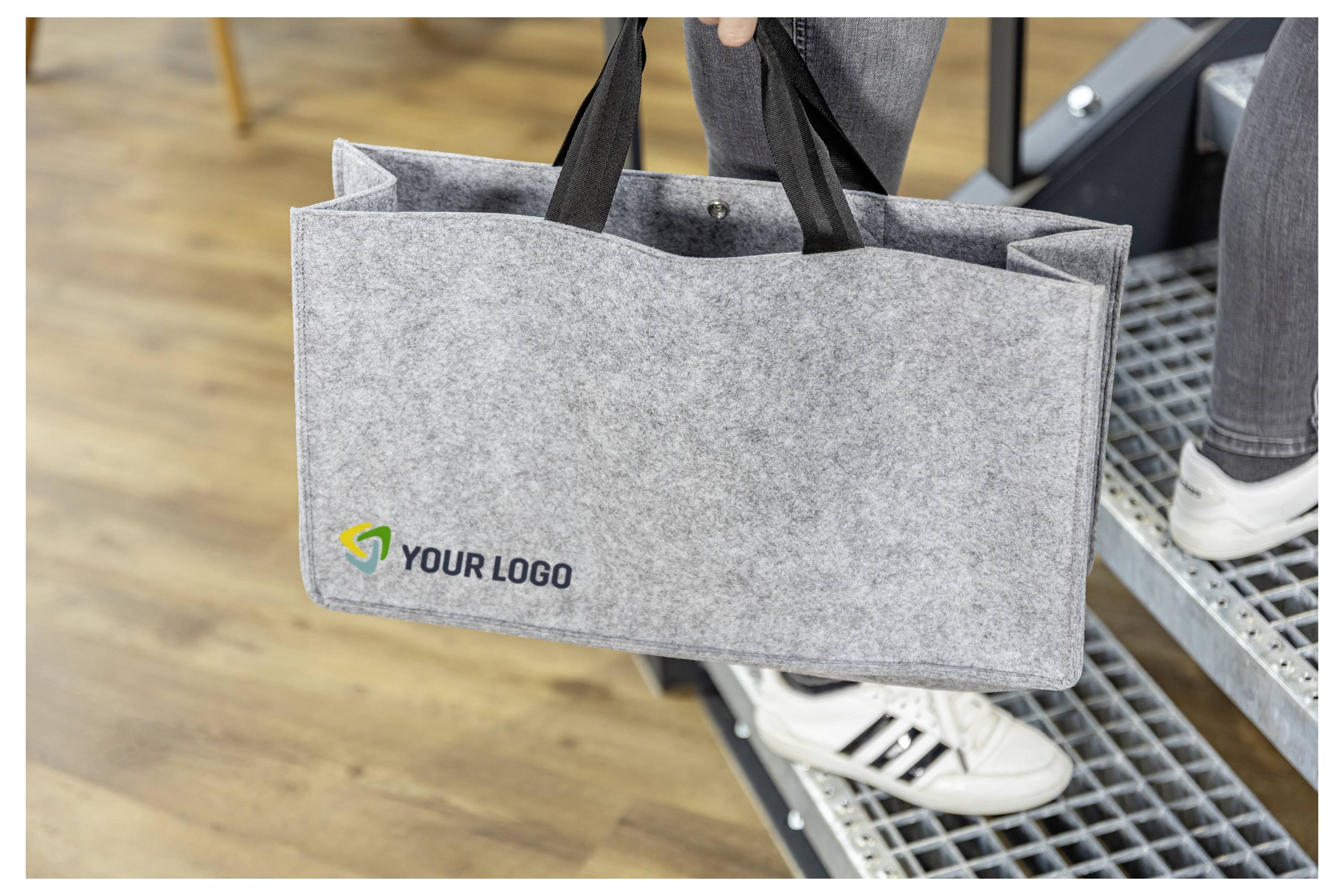 Osoba nesoucí šedou filcovou tašku s potiskem "YOUR LOGO" při chůzi vzhůru po vnitřních schodech.