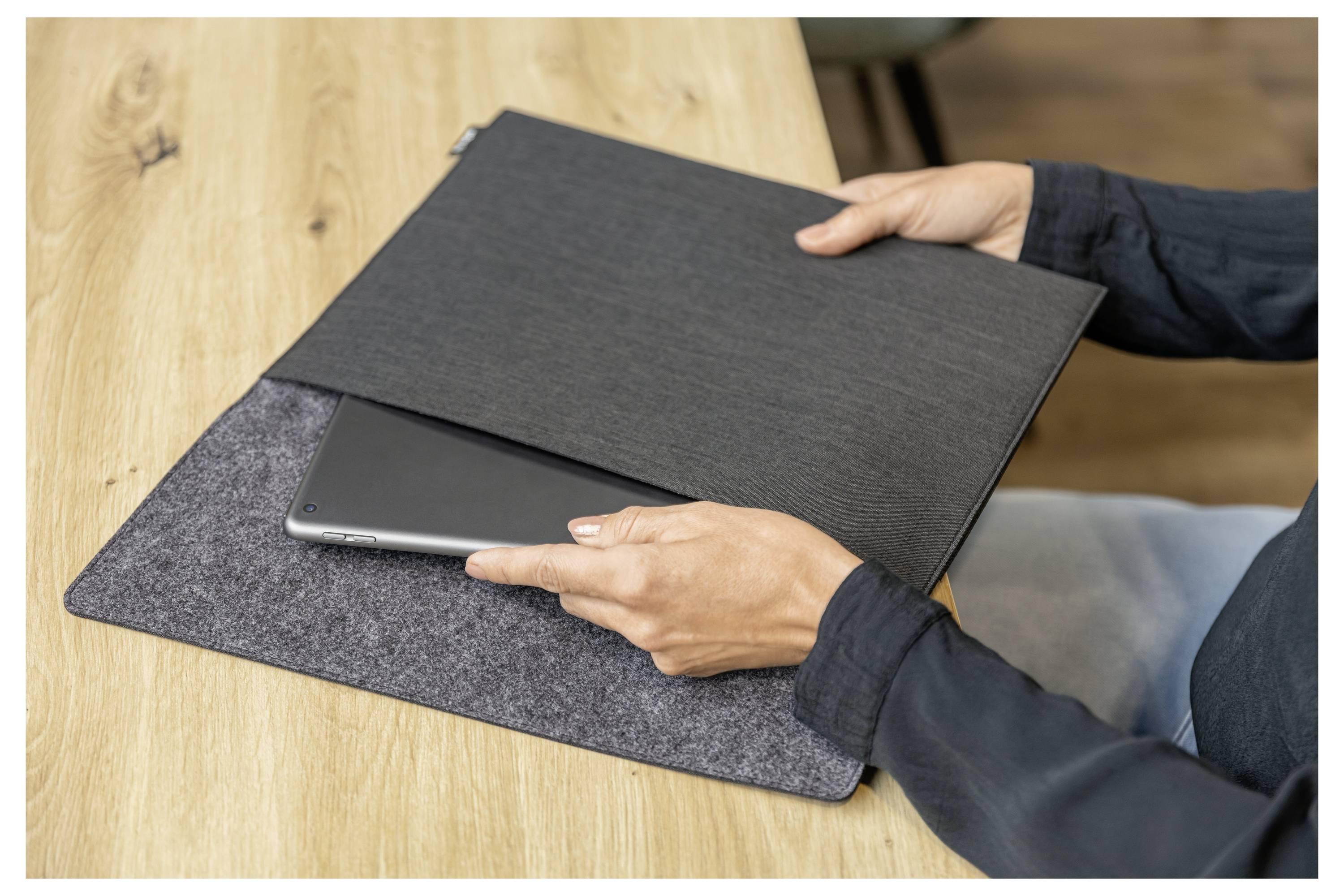 Osoba vkládá notebook do tmavého plstěného pouzdra na dřevěném stole.