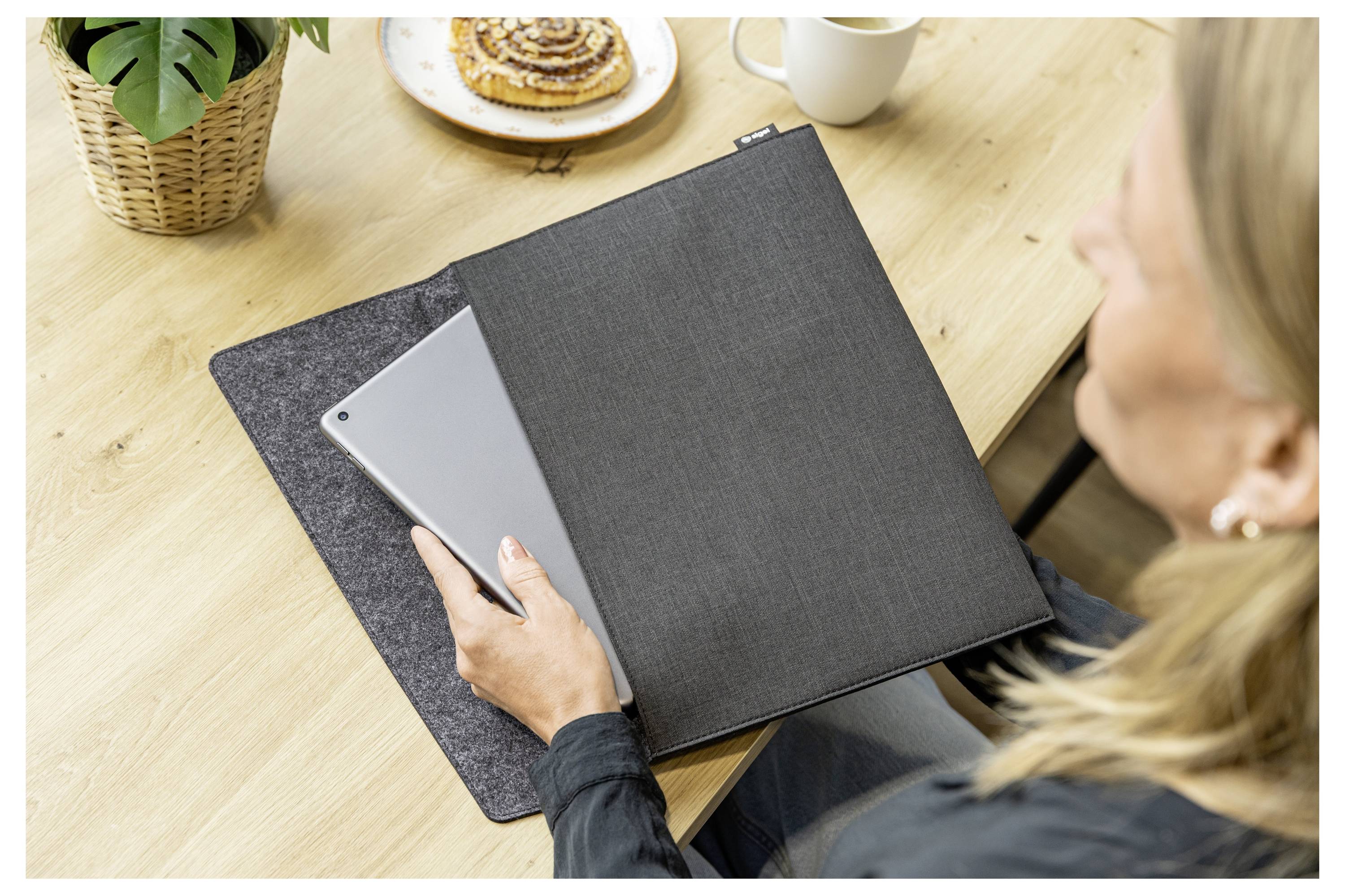 Žena u stolu používá tablet vedle zavřené černé složky, s kávou a syrečkem na stole.