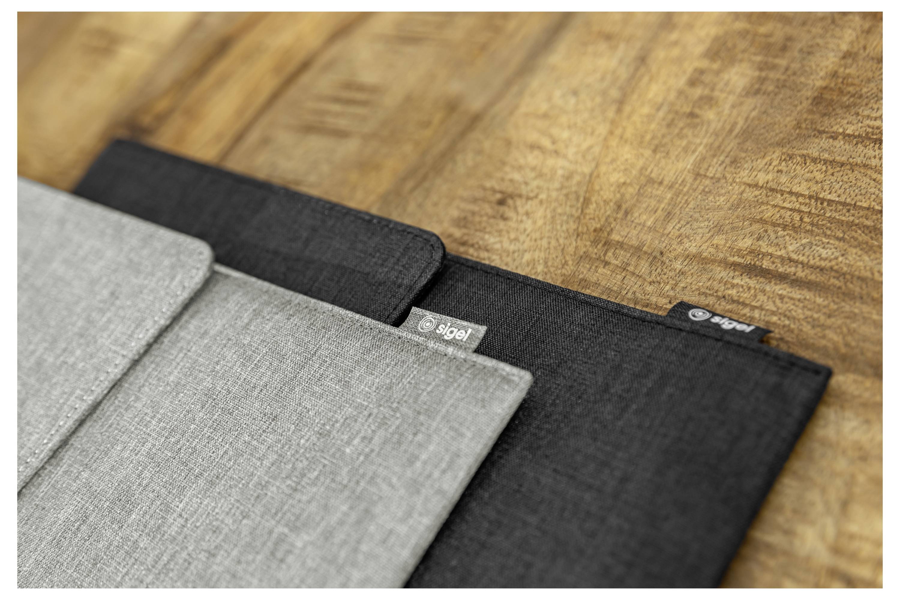 Tři látkové obaly na notebook v šedé a černé barvě jsou naskládány na dřevěné ploše, která ukazuje jejich štíhlý ochranný design a značky výrobce.