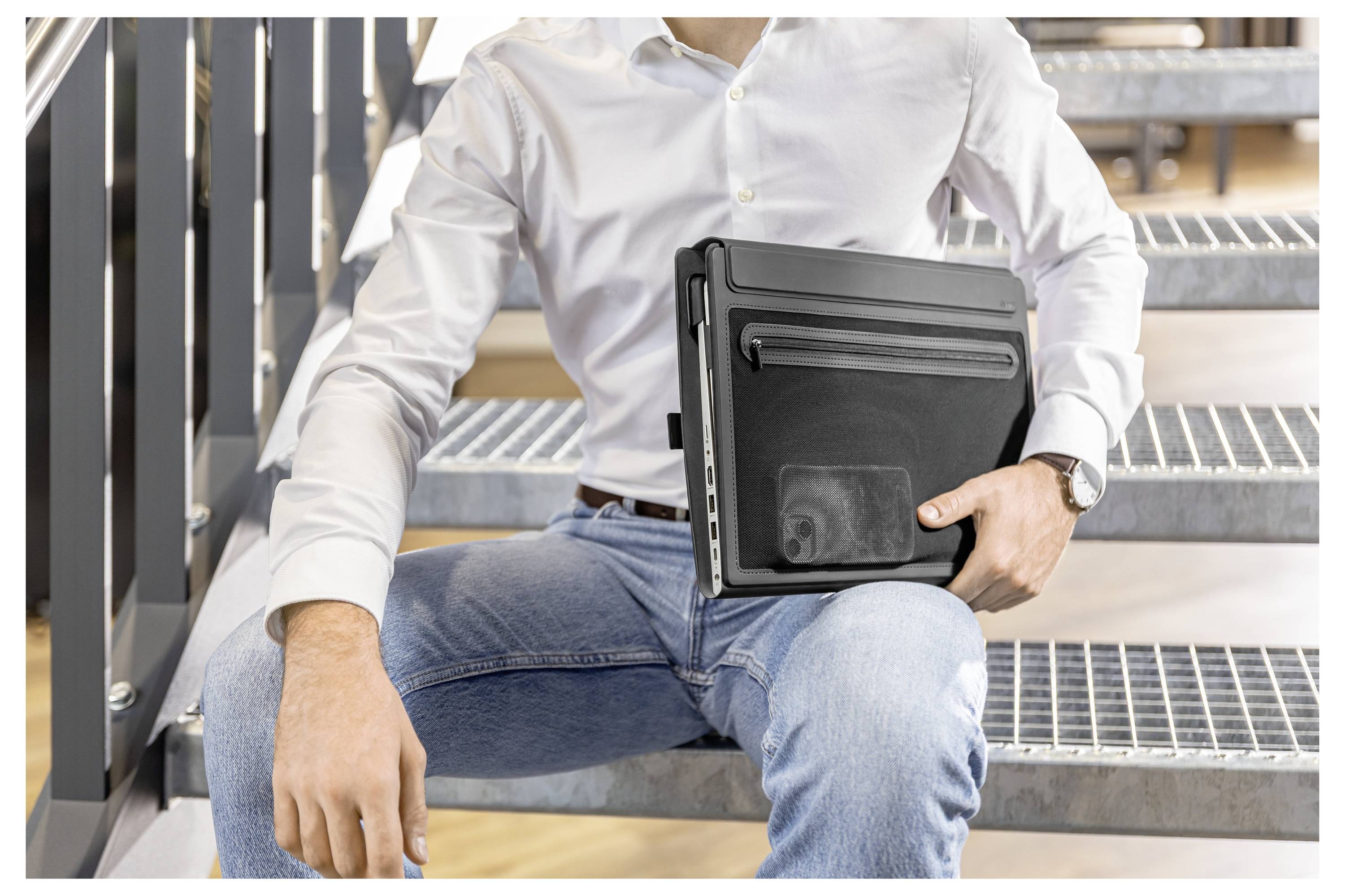 Muž držící zavřený notebook na venkovních kovových schodech, oblečený v bílé košili a džínech.