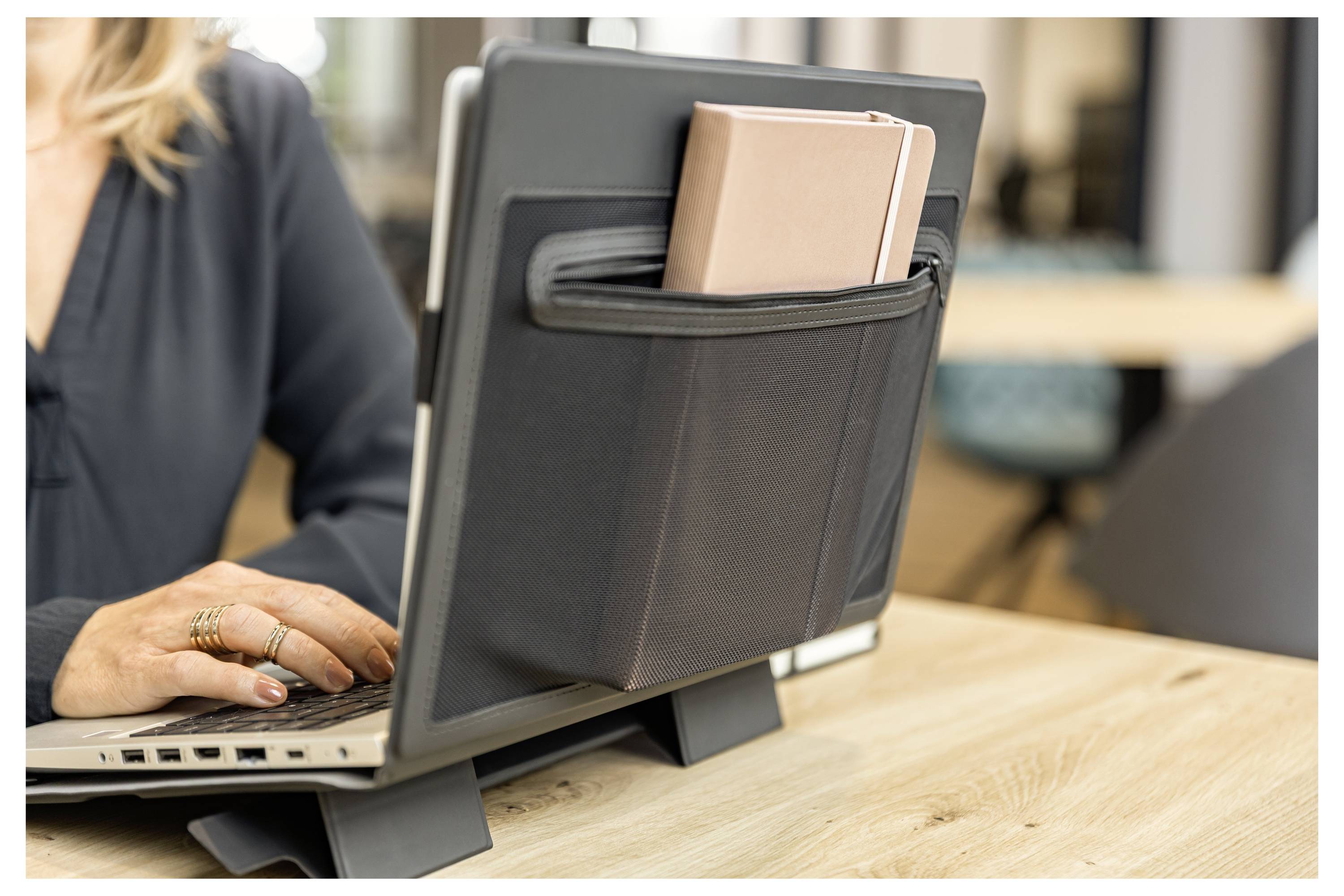 Notebook na stole s organizérem ze sítě připevněným na zadní straně, který drží papíry, zatímco osoba vedle něj píše.