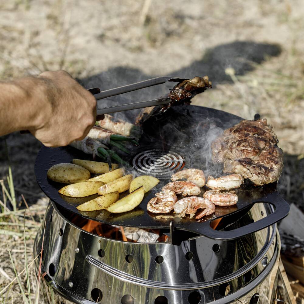 Ruka používá kleště na grilování masa, krevet a brambor na kulatém uhlíkovém grilu venku.