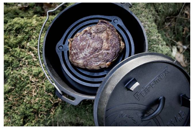 Steak vařící se v litinové holandské peci na venkovním ohni s víkem položeným vedle ní na trávě.