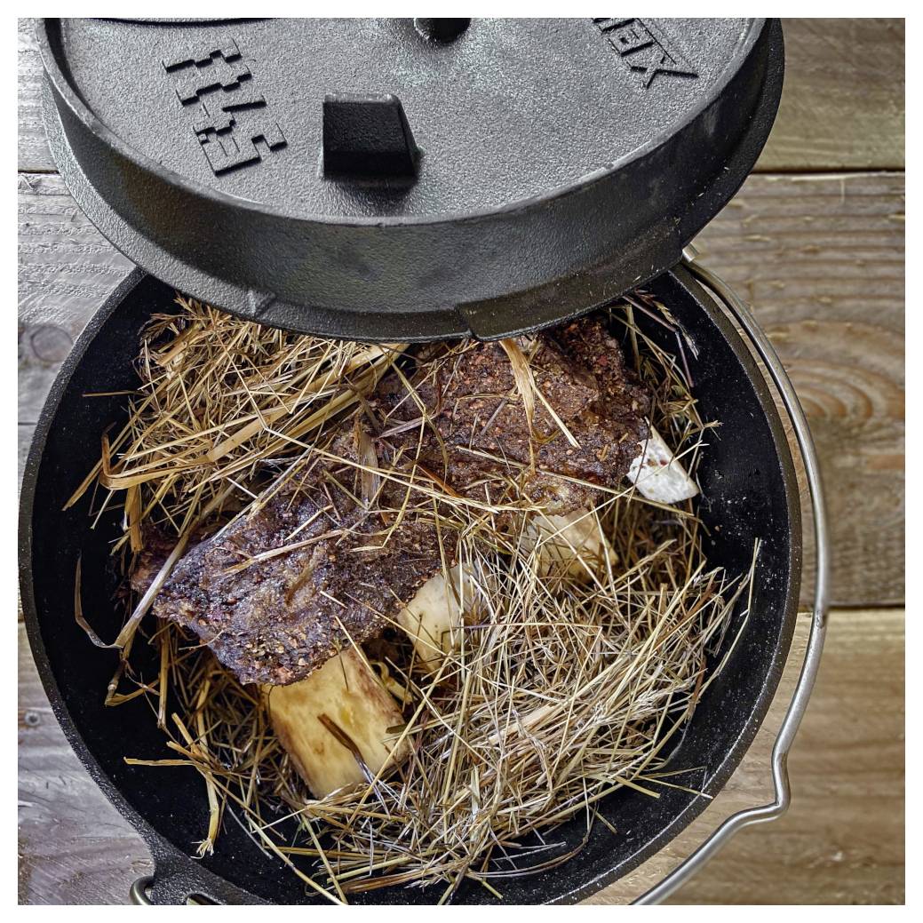 Pohled shora na litinový Dutch oven se zaklopeným víkem, obsahující zhnědlé mясné kosti pokryté slámou na dřevěném povrchu.