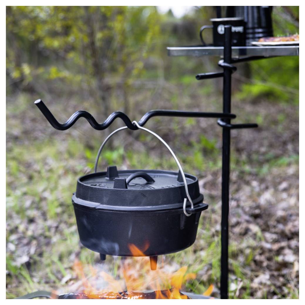 Černý litinový hrnec висí nad táborovým ohněm na trojnožce grilu venku, s viditelnými plameny pod ním.