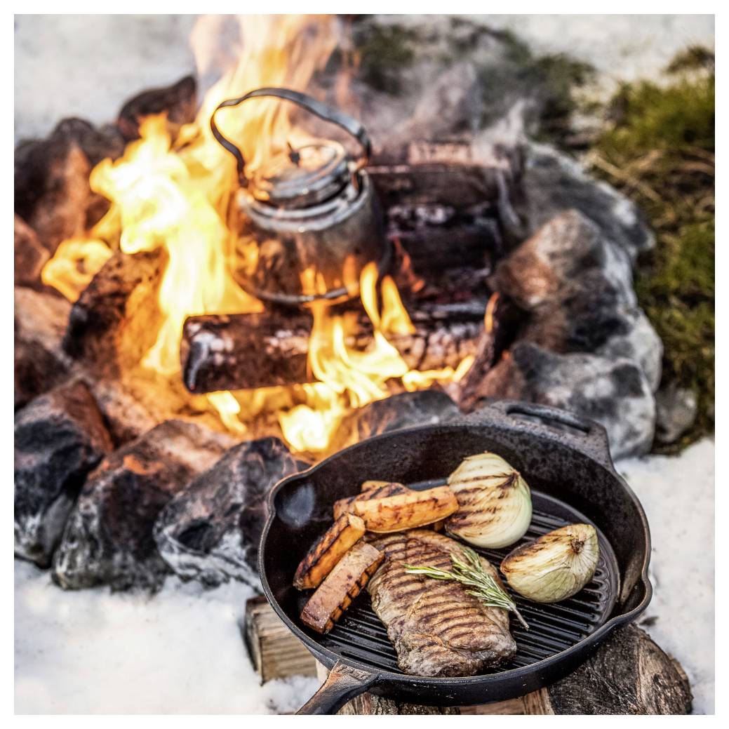 Steak, cibule a sladké brambory se vaří v litinové pánvi nad táborákem, v pozadí se na plamenech ohřívá konvice.